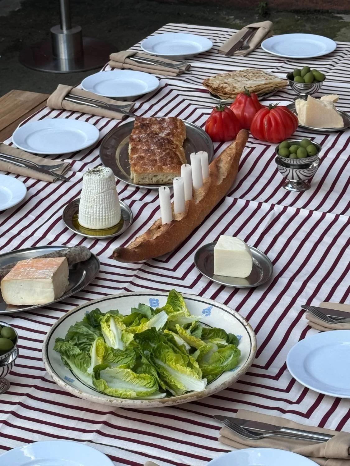 A table set with various foods including lettuce, cheeses, bread, olives, tomatoes, and a cake, with plates, cutlery, and napkins arranged for a meal.