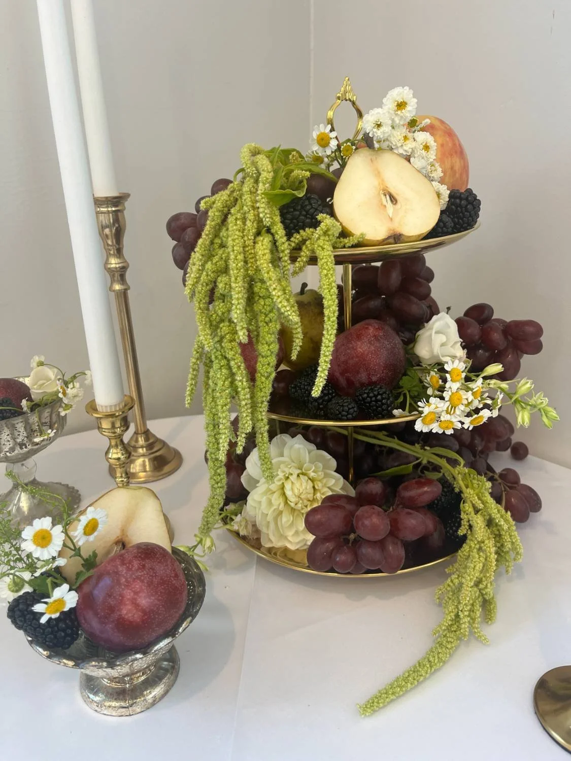 Elegant display of apples, grapes, blackberries, and daisies arranged on a tiered gold stand and nearby dishes, with candles and silverware in the background.