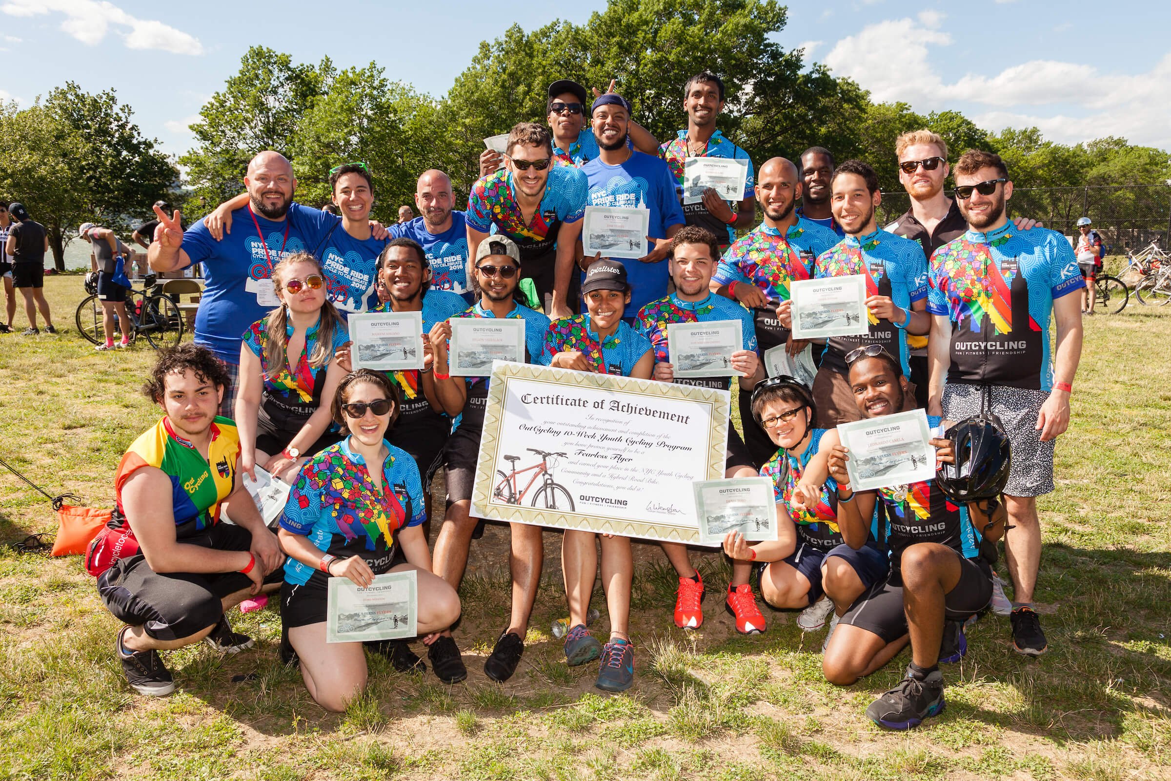 Group of people holding certificates and a large certificate, posing outdoors on a grassy field with trees in the background on a sunny day.