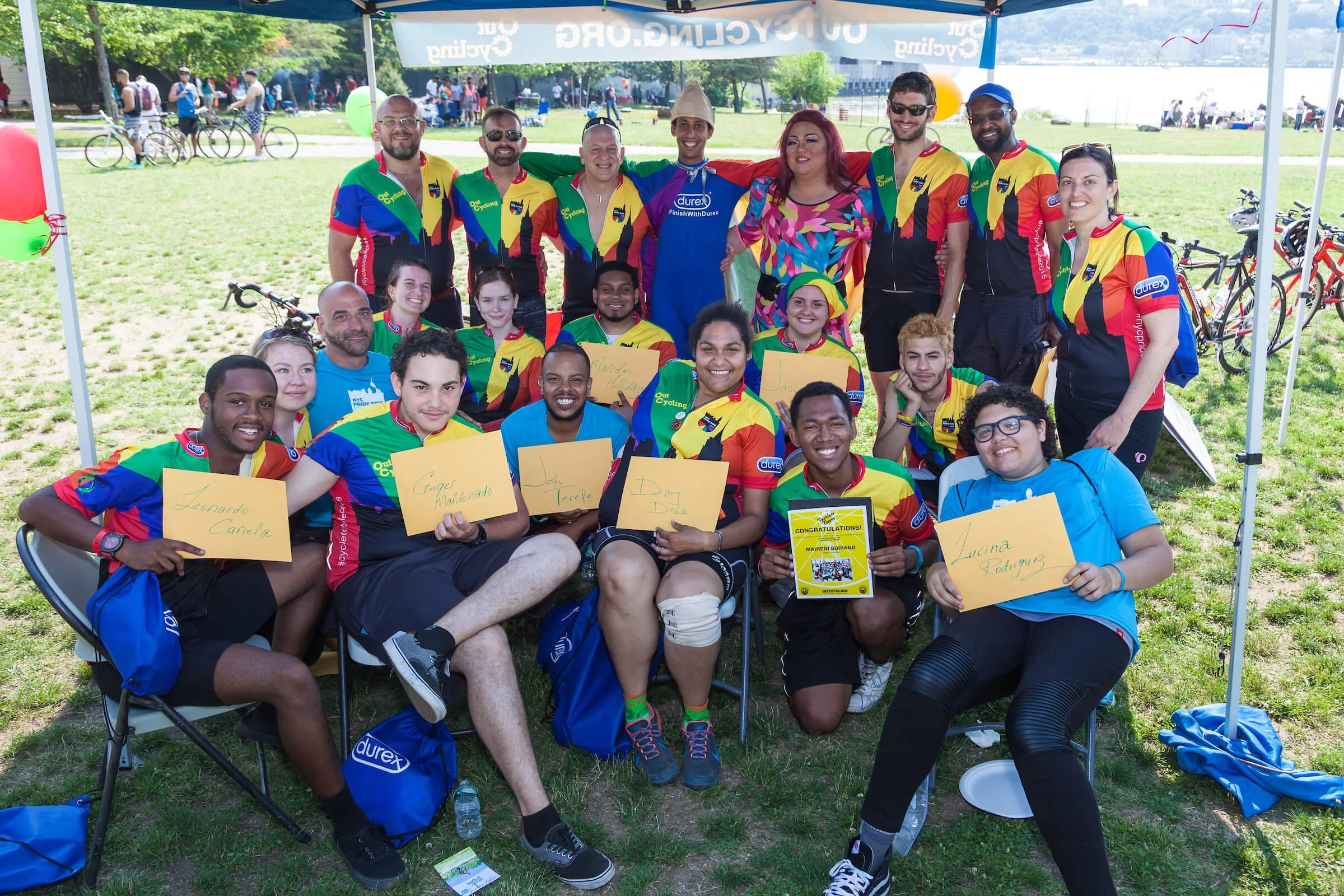 Group of smiling people at an outdoor event, some sitting with yellow signs displaying names, others standing in colorful cycling jerseys, with a park and lake in the background.