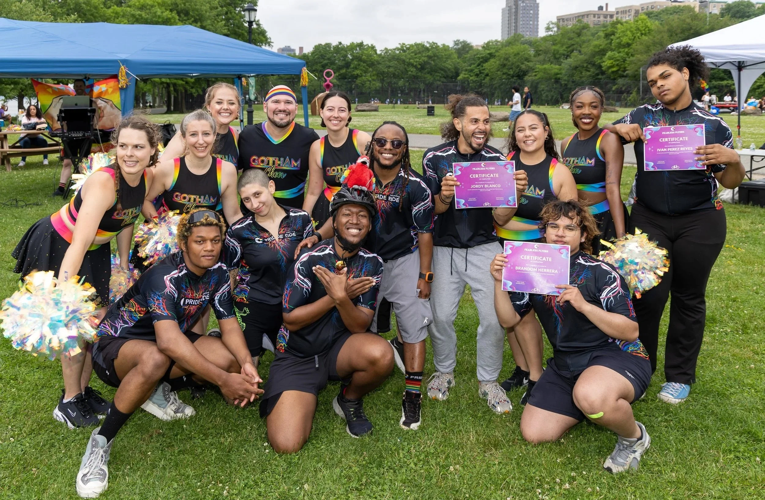 A group of people outdoors in a park, wearing matching cycling team outfits, some holding certificates, and posing for a team photo under tents with trees and city buildings in the background.