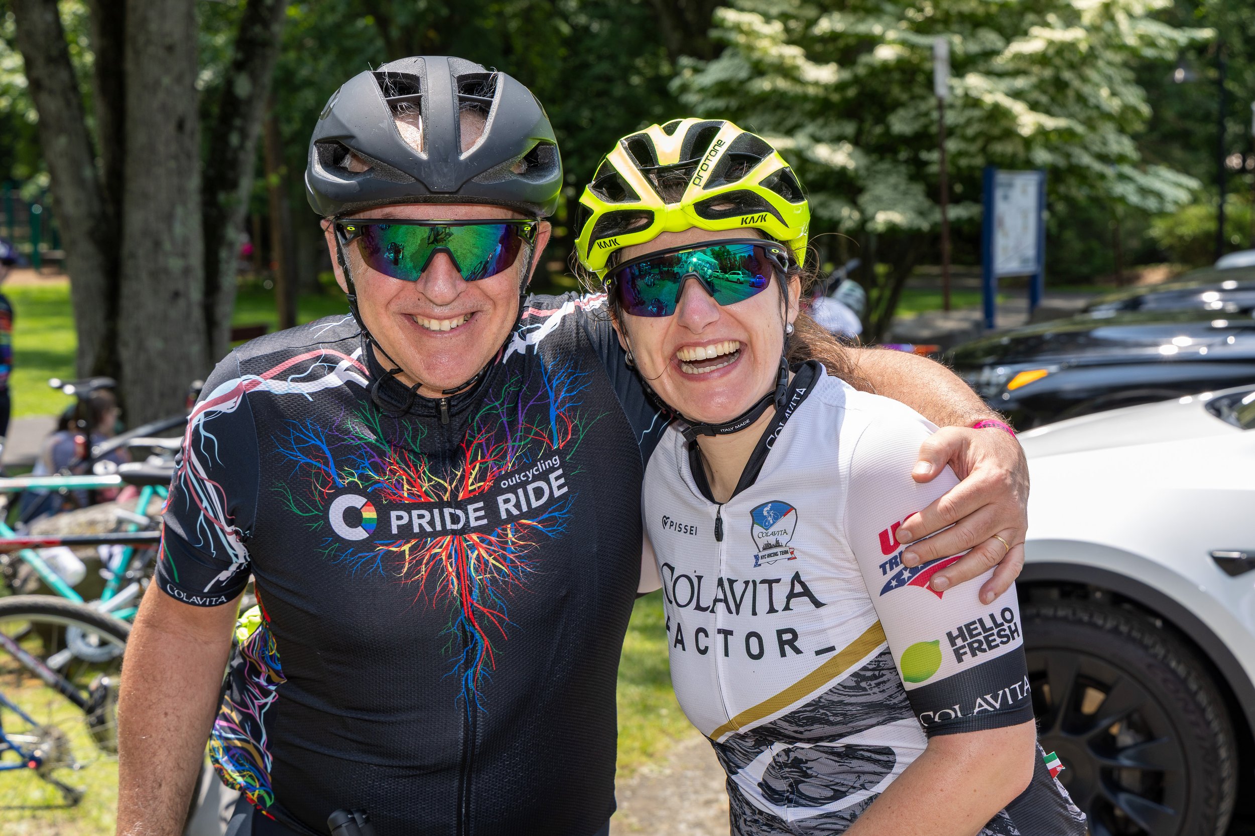 Two cyclists wearing helmets and sunglasses smiling and hugging each other outdoors near a parking lot with trees and cars in the background.