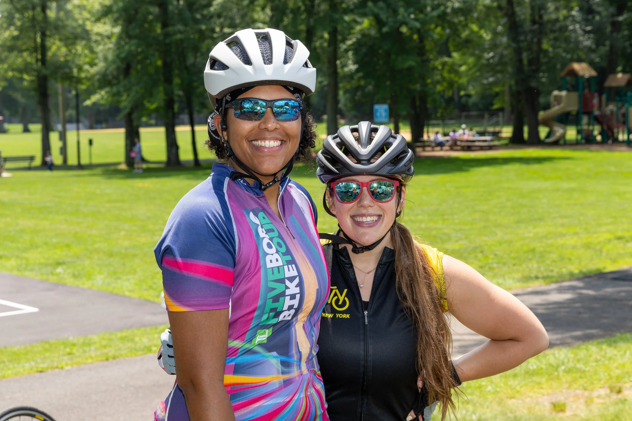 Two women wearing cycling helmets and sunglasses smiling in a park with trees and playground equipment in the background.