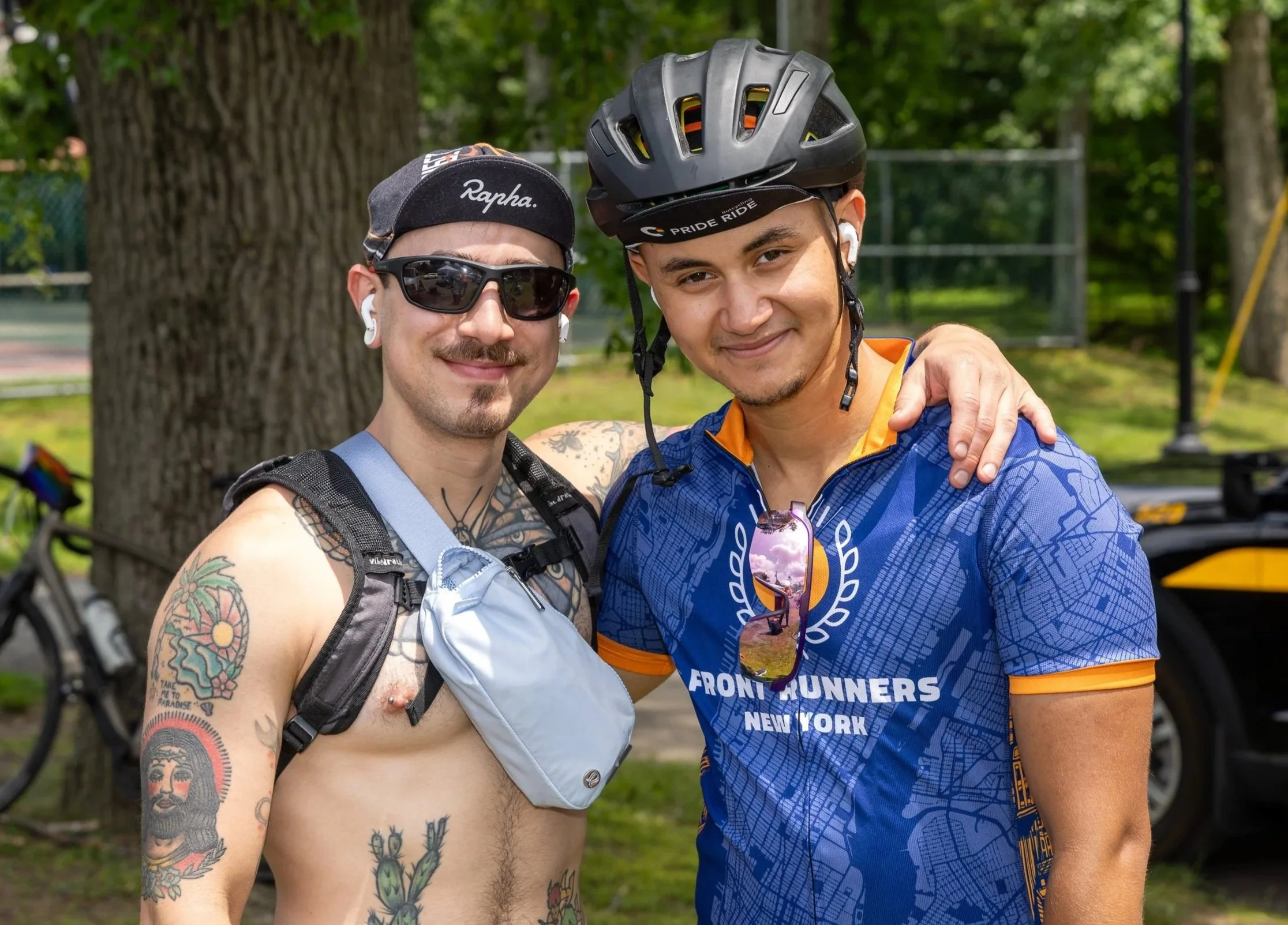 Two men outdoors, one shirtless with tattoos and sunglasses, the other in a blue cycling jersey and helmet, smiling and posing together.