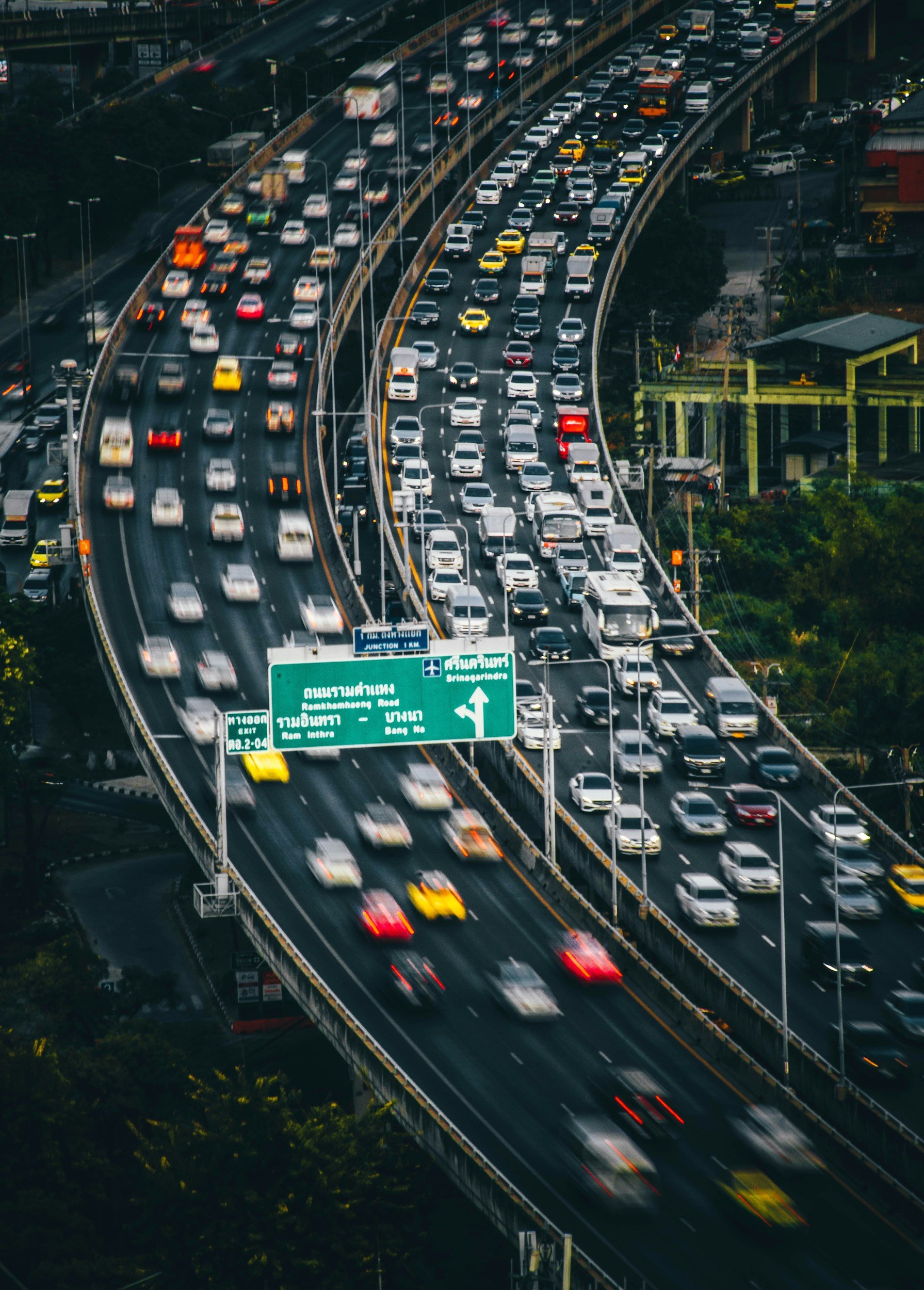 A busy elevated highway filled with cars, trucks, and taxis moving in traffic, with green overhead signs displaying directions in Thai and English.