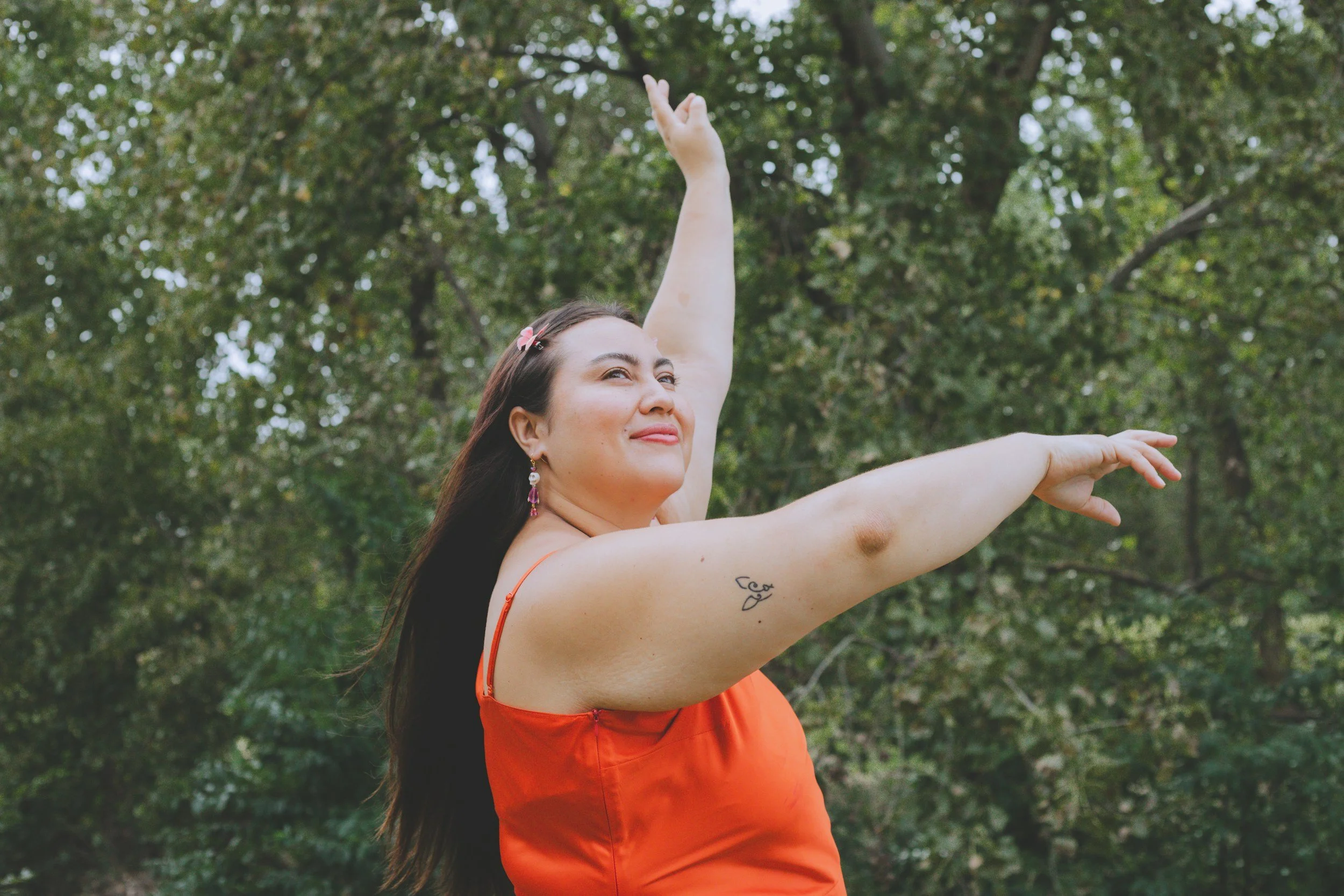 A woman with long brown hair, wearing an orange dress and pink earrings, is outside with her arms raised in a dance or yoga pose. She is smiling slightly and standing in front of a background of green trees.