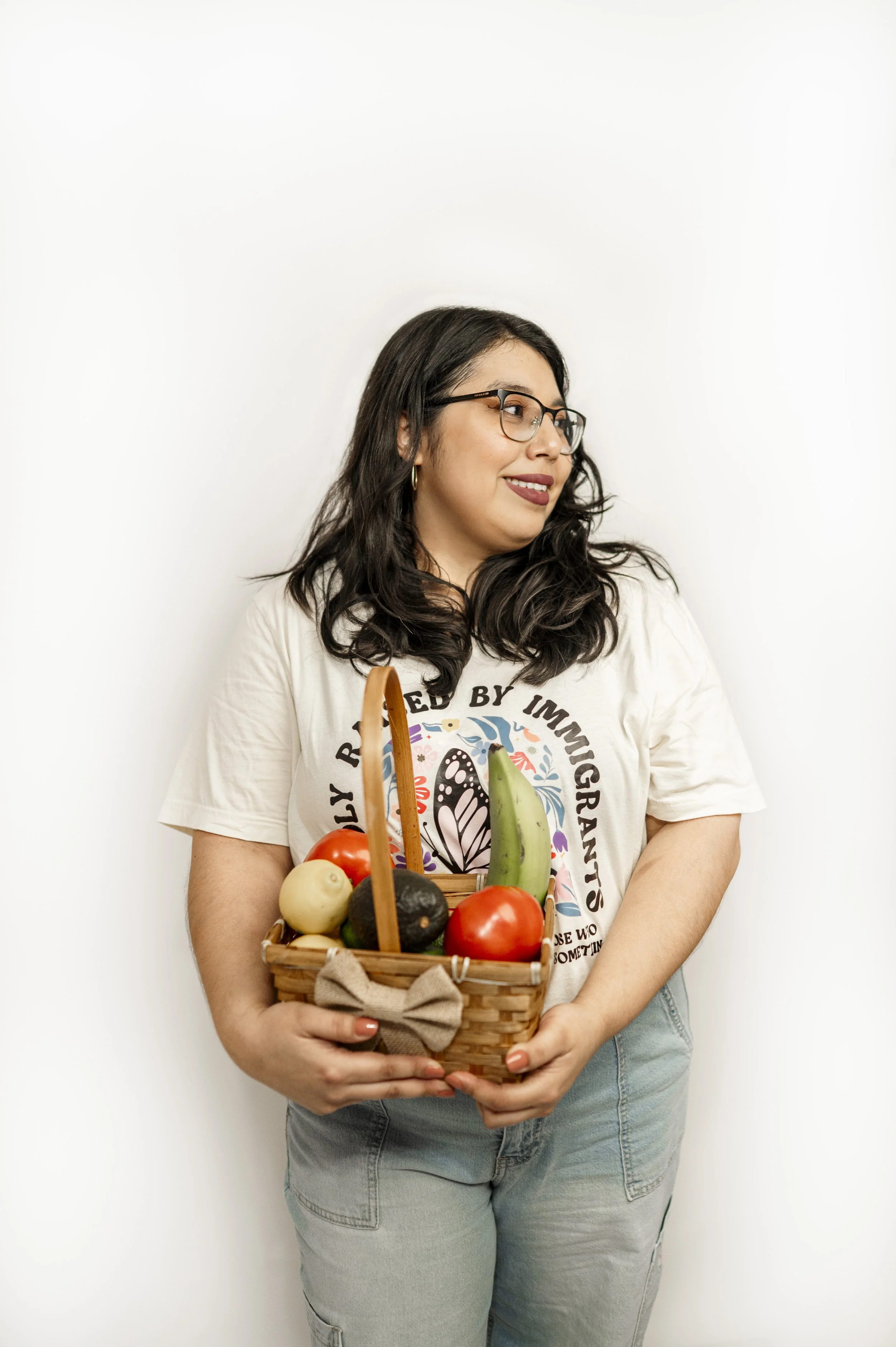 Pictured is Ashley, a woman with long dark hair, glasses, and hoop earrings holding a basket filled with various vegetables, standing against a plain white wall.