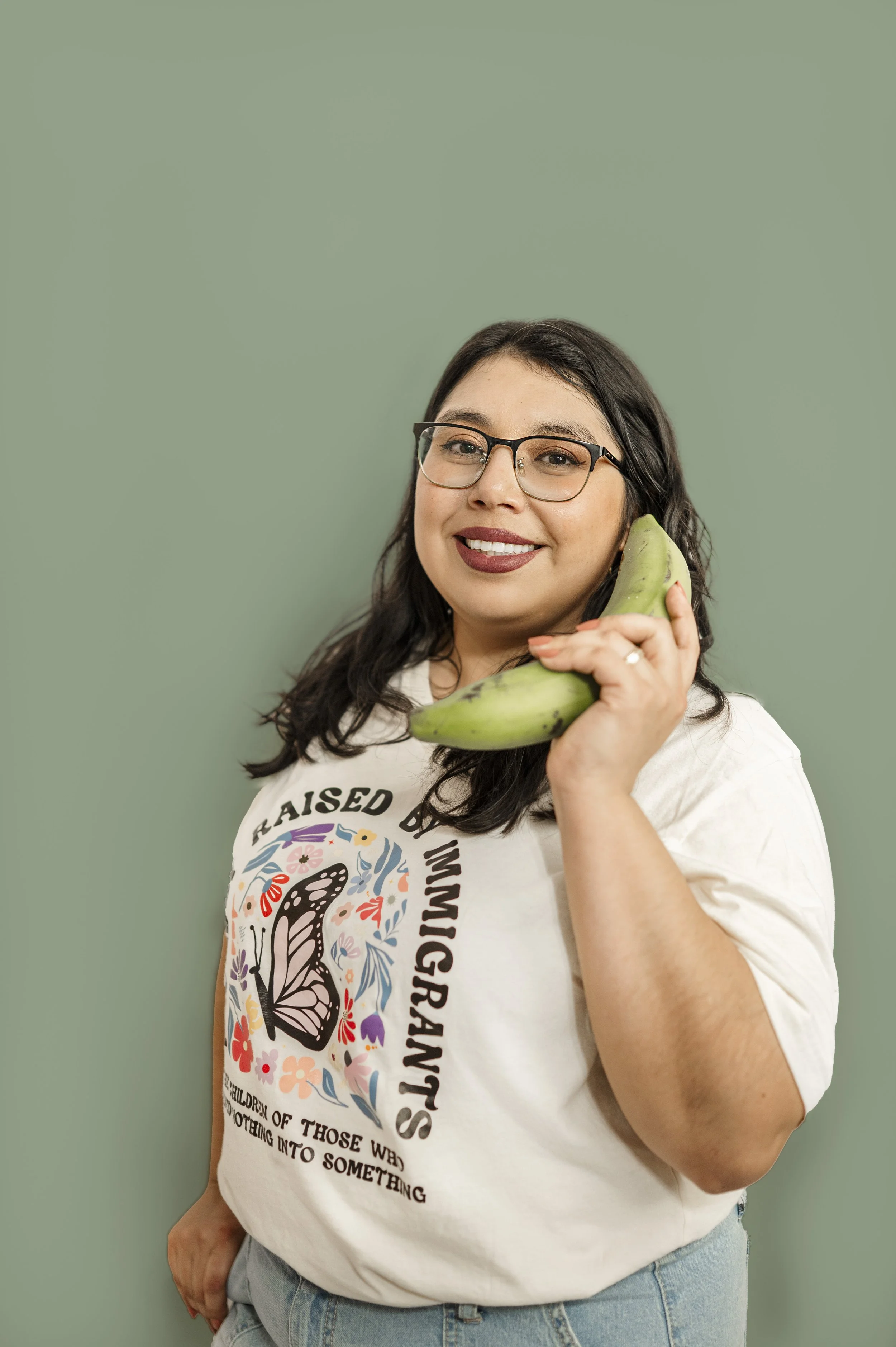 Pictured is Ashley, a woman with dark hair, glasses, and a wide smile, holding a green plantain near her face like a telephone, standing against a muted green background.