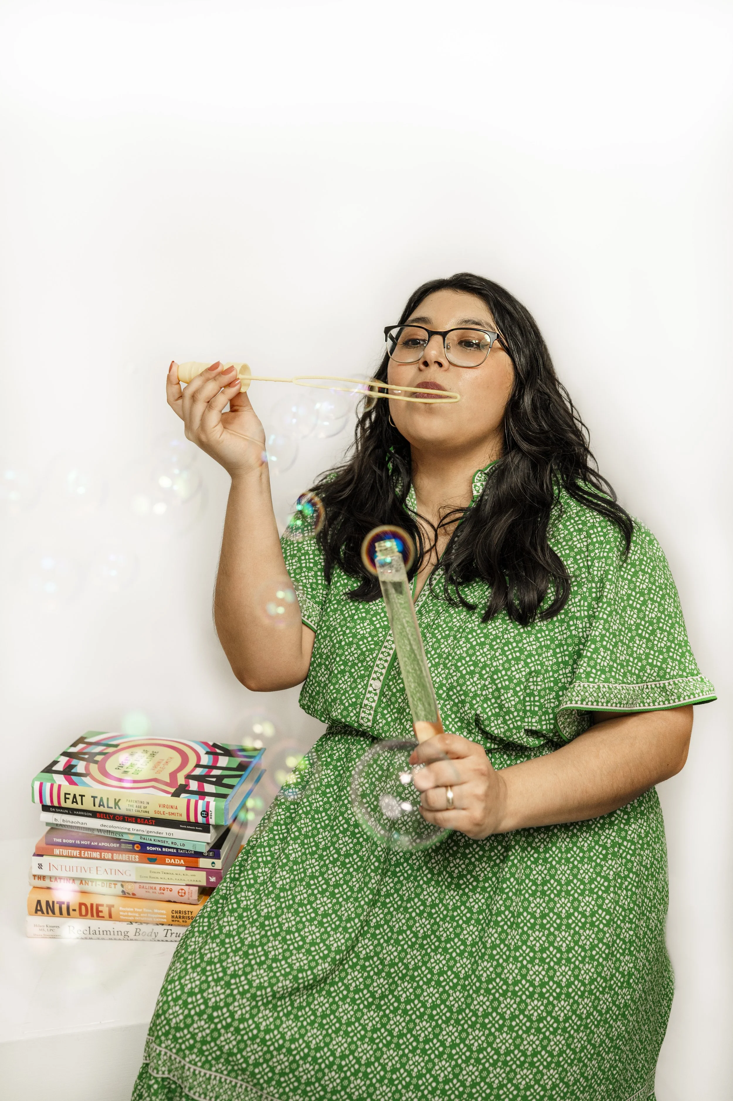 Pictured is Ashley, a woman with long dark hair and glasses sitting in front of a white background, blowing bubbles with a bubble wand. She wears a green patterned dress. A pile of books on inclusive health topics is on the table beside her.