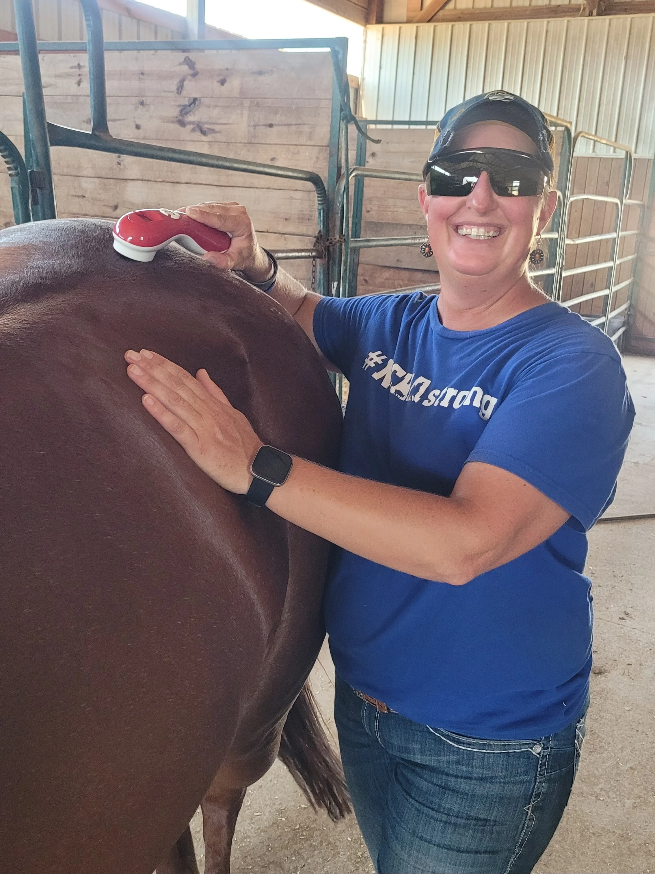 A woman with sunglasses and a blue shirt stands next to a horse, smiling, while holding a cold laser on the horse's body.