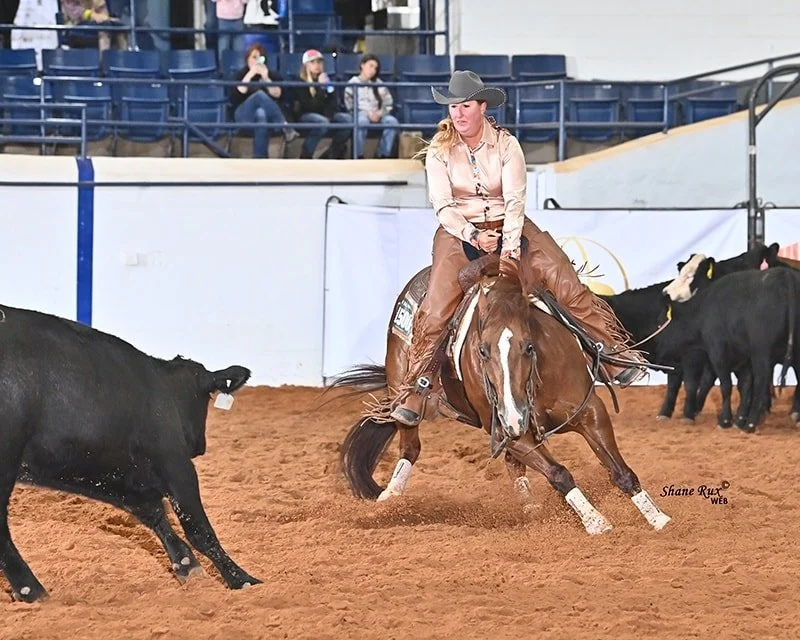 A woman in western attire riding a cutting horse in an indoor arena with spectators watching from the stands.