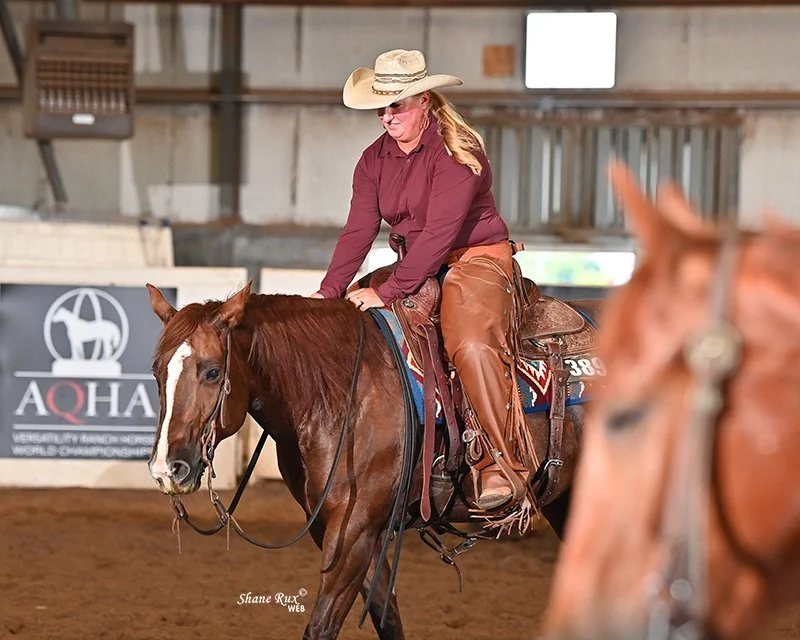 A woman wearing a cowboy hat and maroon shirt riding a brown horse inside an indoor riding arena.