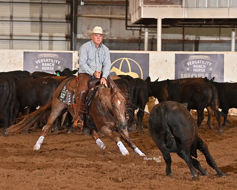 A cowboy riding a red roan horse, wearing a cowboy hat, is participating in a cutting show in an indoor arena.