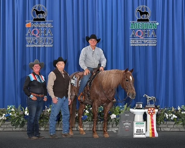 Three people, two women and one man, standing in front of a blue curtain at an AQHA horse show. The man is sitting on a brown horse wearing western attire and a cowboy hat. The women are also dressed in western-style clothing and cowboy hats. There are flowers and awards displayed on the floor, including a ribbon and a sign indicating a reserve champion title.