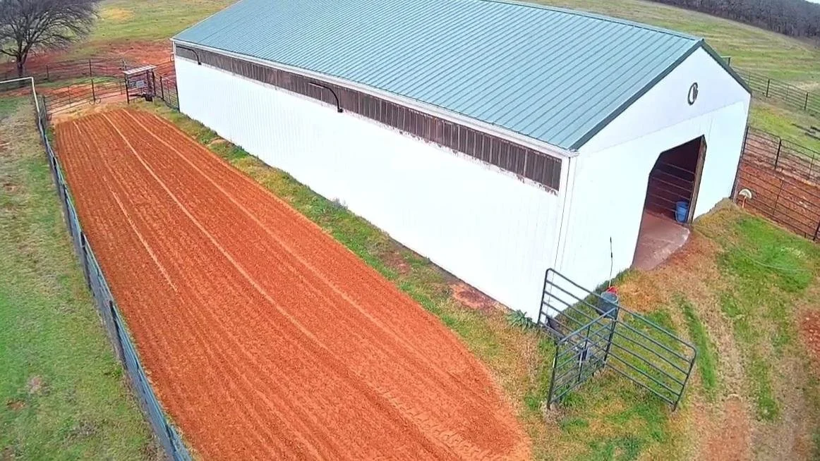A large white barn with a light blue metal roof, adjacent to a freshly tilled red soil field, enclosed by black metal fencing, with a person near the entrance.