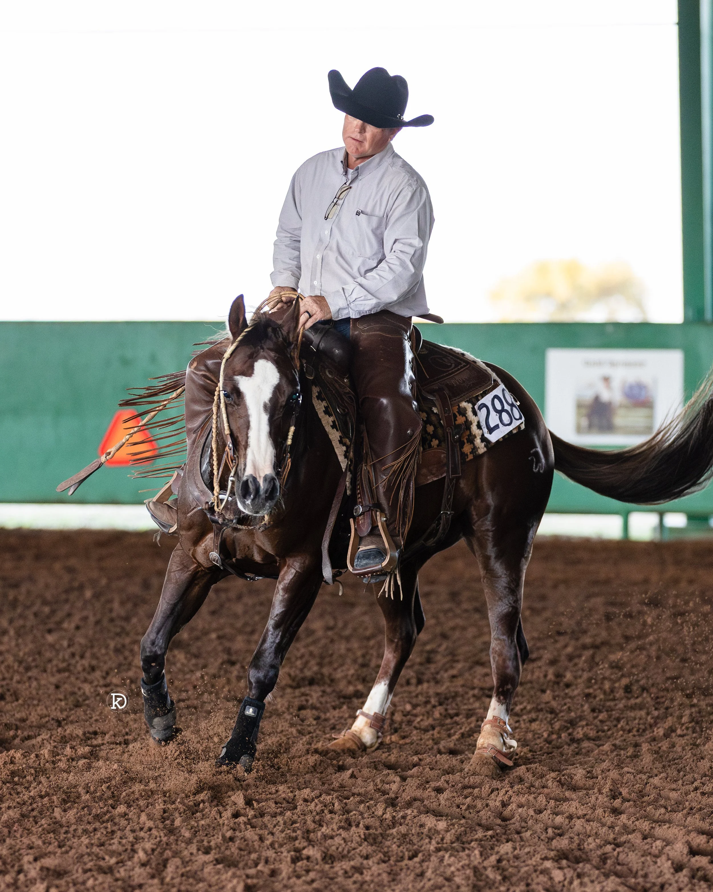 Man in a white shirt and black cowboy hat riding a brown and white horse inside an indoor riding arena.
