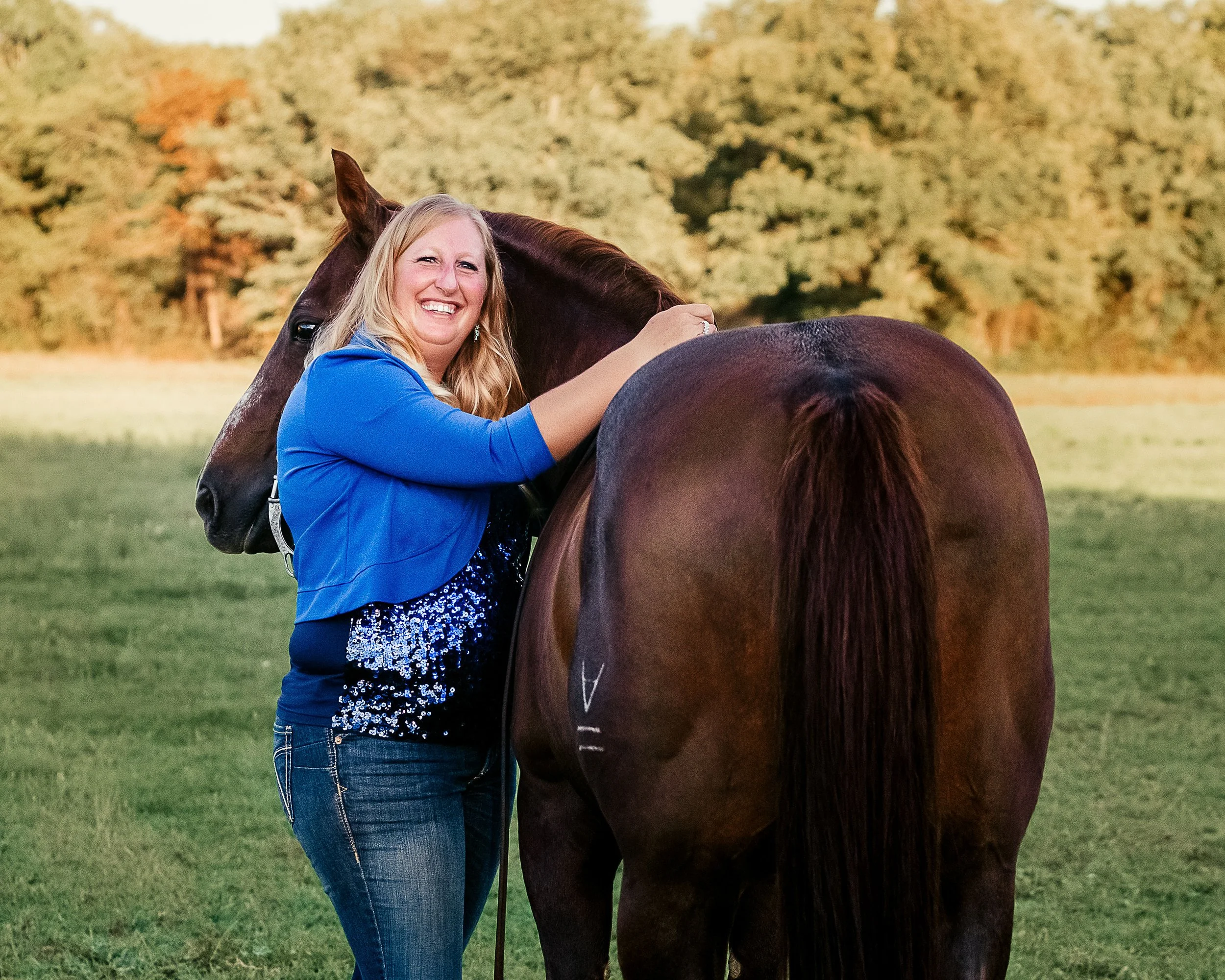 A woman in a blue jacket and jeans standing outdoors, smiling and leaning on a brown horse with a background of trees and a grassy field.