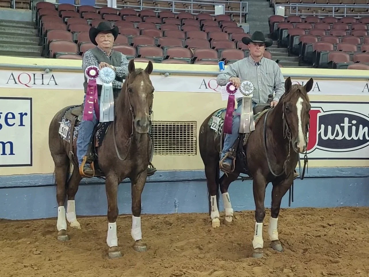 Two riders on horseback holding ribbons and trophies inside the Jim Norick arena with empty bleachers.