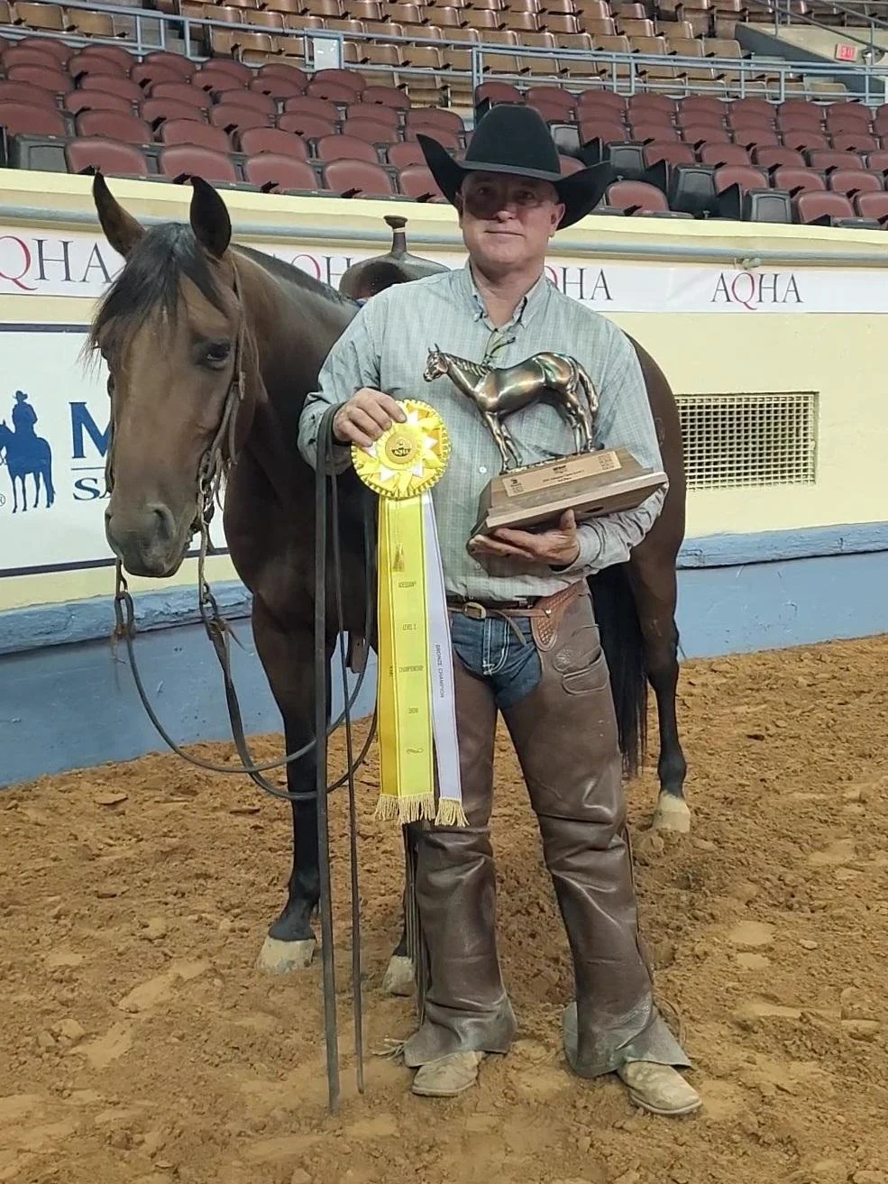 A man wearing a cowboy hat and jeans holding a trophy and ribbon standing next to a brown horse in an arena with empty red seats in the background.