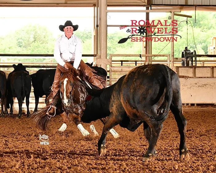 A cowgirl riding a horse in a cutting competition, with a black cow in front of the horse. Several other cattle are visible in the background at an indoor livestock arena, with text indicating a 2024 Romal's & Rowels Showdown event.