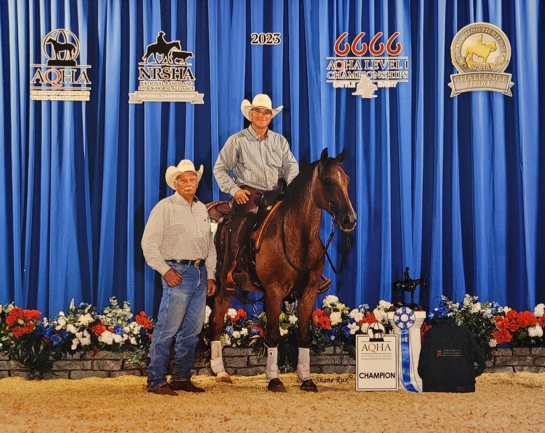 Two men in cowboy hats and jeans, one on a horse, standing in front of a blue curtain and award banners at a horse competition. The horse is a red roan, wearing leg wraps, and there's a champion sign and ribbons on the ground.