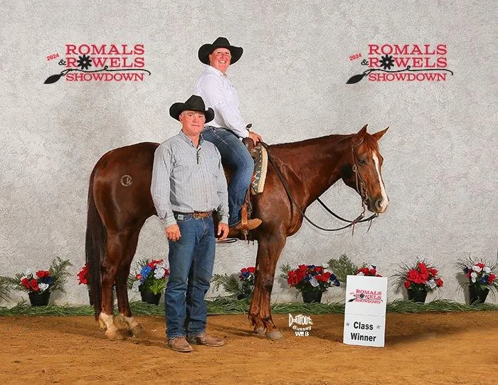 A woman in a white shirt and black cowboy hat sits on a brown horse, with a man in a gray plaid shirt and black cowboy hat standing beside them. They are in an indoor arena with red, white, and blue flower arrangements and a sign that reads 'Class Winner.' The background features a banner with the text 'Romals & Rowels Showdown 2024.'