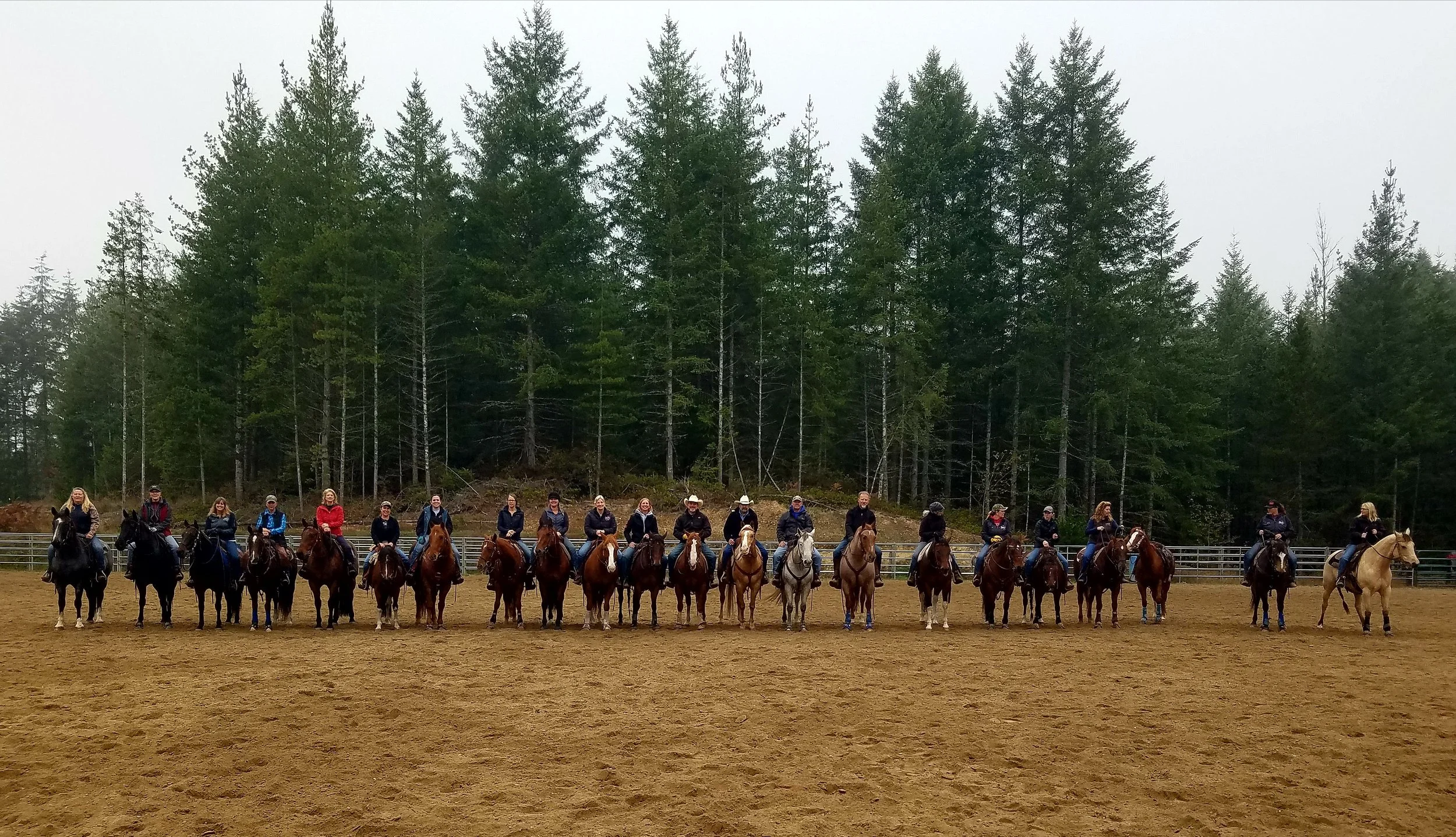 A line of twenty-one people on horseback in an outdoor riding arena with a forest of tall pine trees in the background.