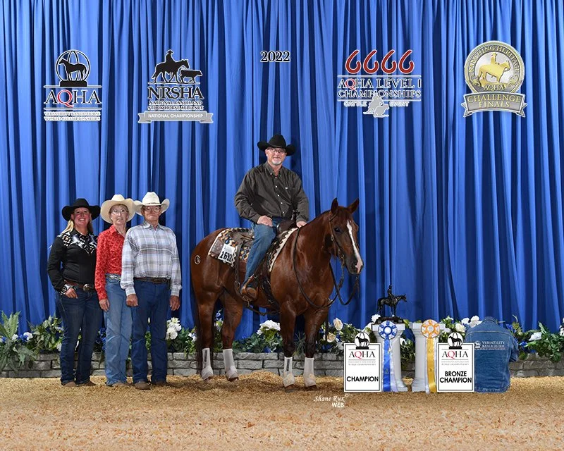 A man sitting on a horse with three people standing beside them in front of a blue curtain at a AQHA World Show, with banners and trophies on the ground indicating a championship win.