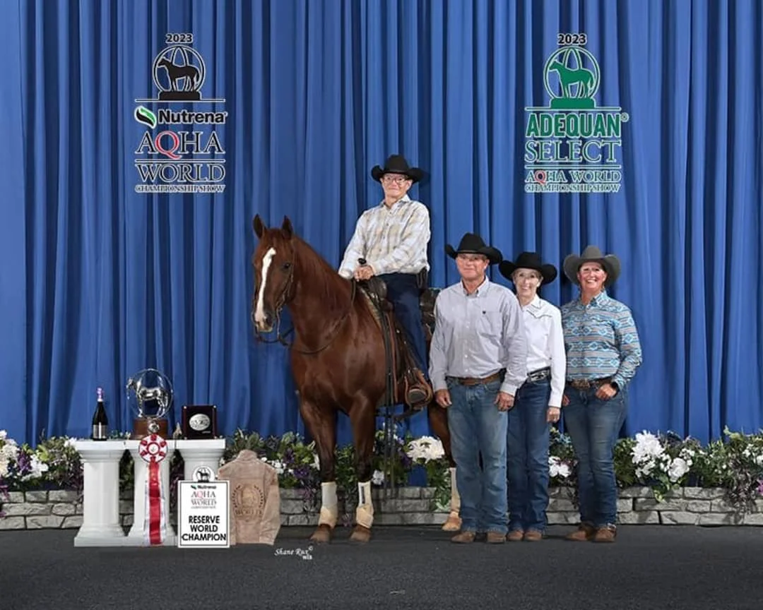 A man on a horse, winning a reserve world championship, with three other people standing beside him, all wearing cowboy hats, in front of a blue curtain with logos and floral decorations.