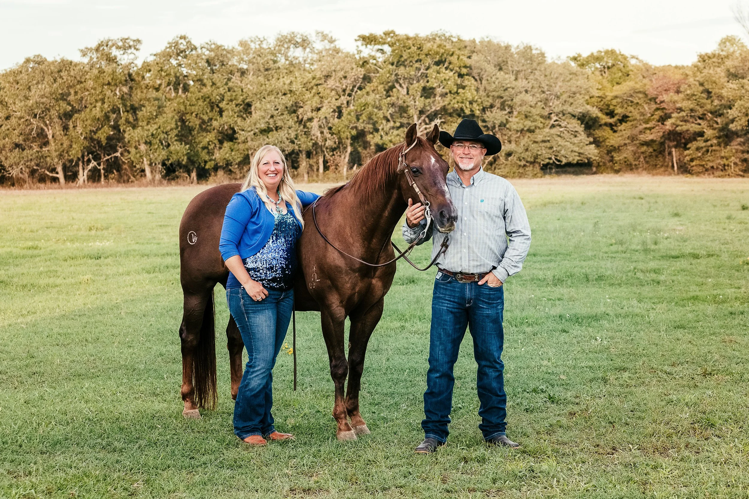 A smiling woman and a man standing beside a brown horse in a grassy field with trees in the background. The woman is wearing a blue jacket and jeans, and the man is wearing a cowboy hat, a light shirt, and jeans. The woman is holding the horse's reins while the man holds the horse's bridle.