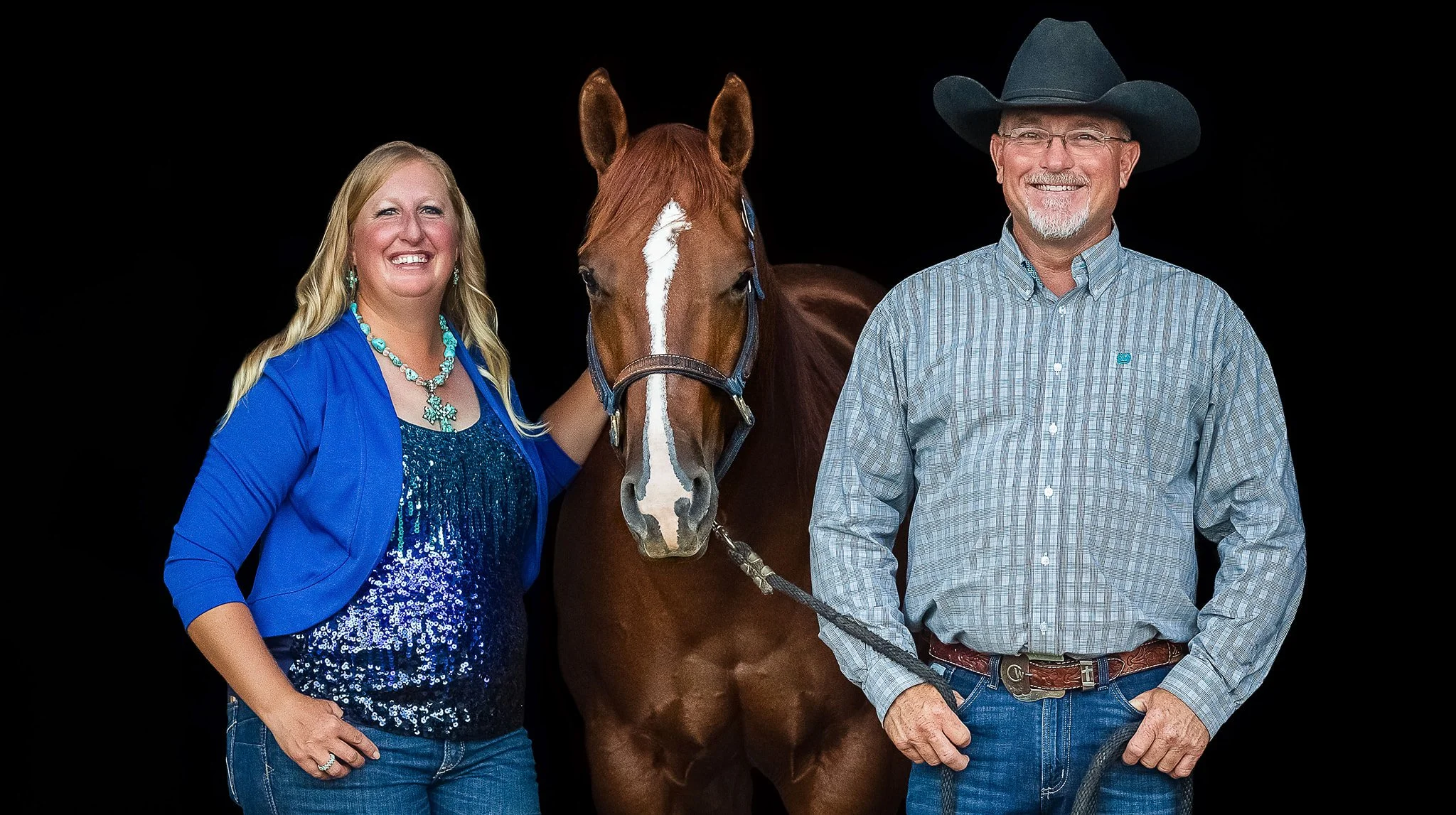 A woman and a man stand with a brown horse against a black background. The woman, wearing a blue jacket and blue sequined top, has long blonde hair and is smiling. She has her hand on the horse's neck. The man, wearing a cowboy hat, glasses, a gray checkered shirt, and jeans, is smiling with his hand near his pocket.