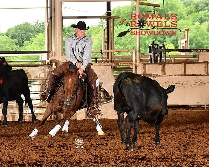 A cowboy on horseback competing in a cutting horse competition inside an indoor arena with a banner reading 'Romels & Rowels Showdown'. The arena has a dirt floor and open sides with greenery outside.