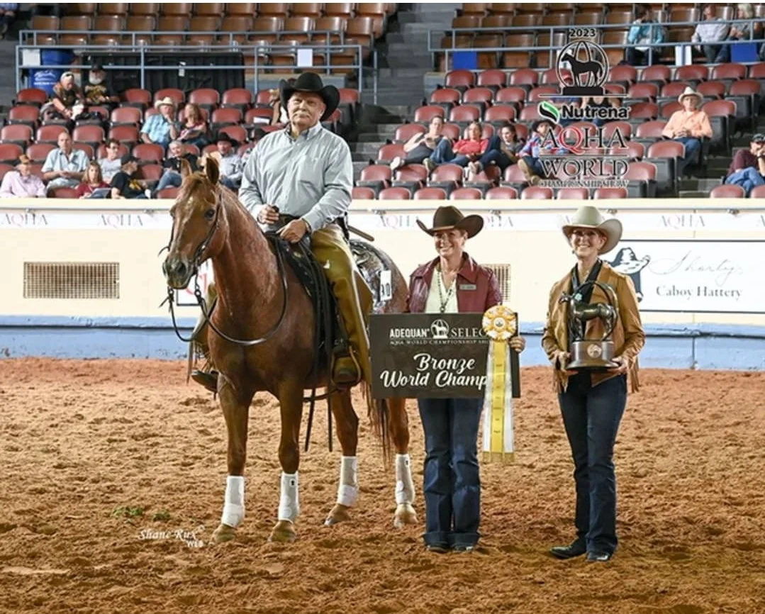 Man on horseback receiving awards at the AQHA World Show event, with two women holding a 'Bronze World Champion' sign and a trophy, in an indoor arena with audience in the background.