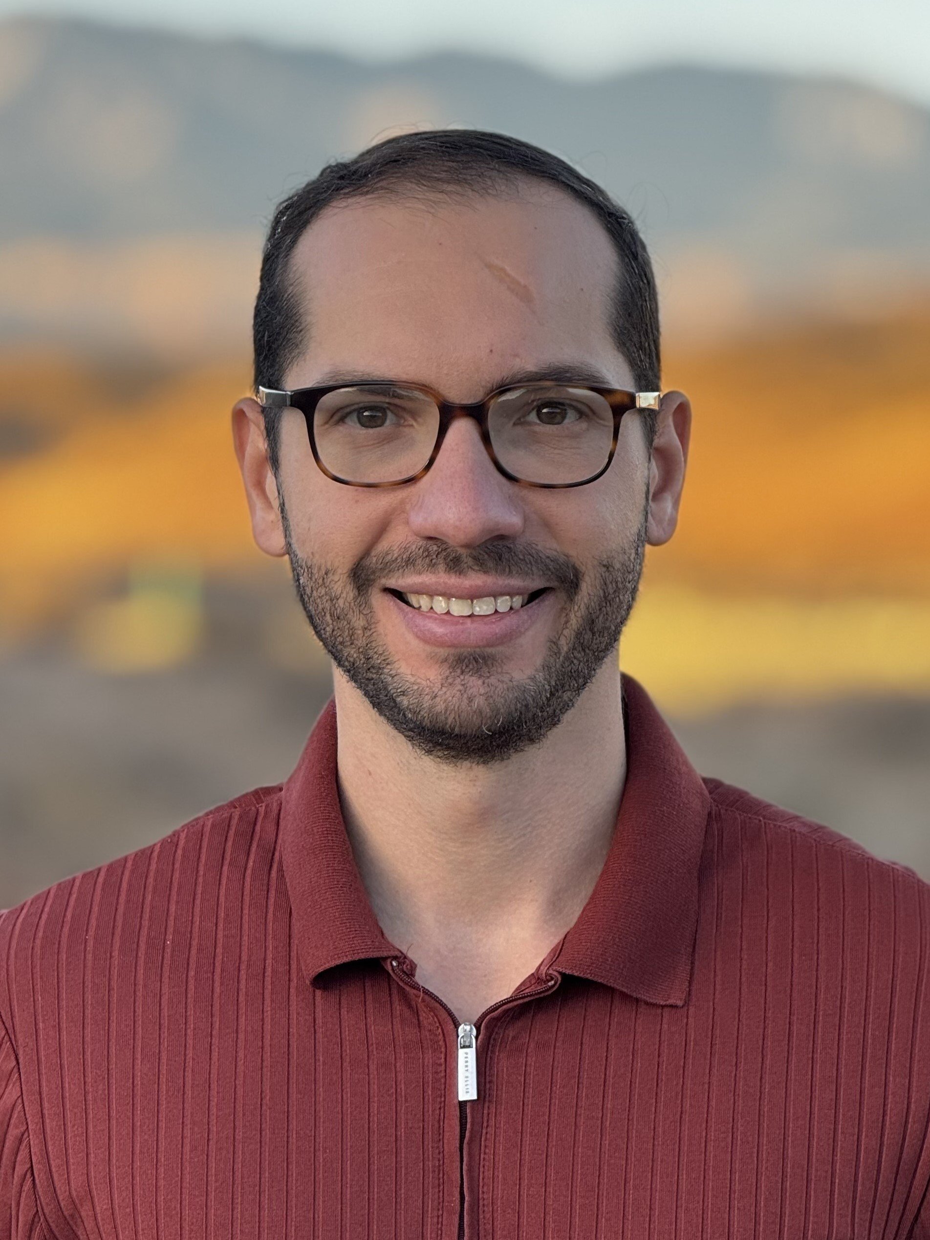 A smiling man with glasses and a beard, wearing a maroon zip-up shirt, outdoors with a blurred background of colorful trees and a clear sky.