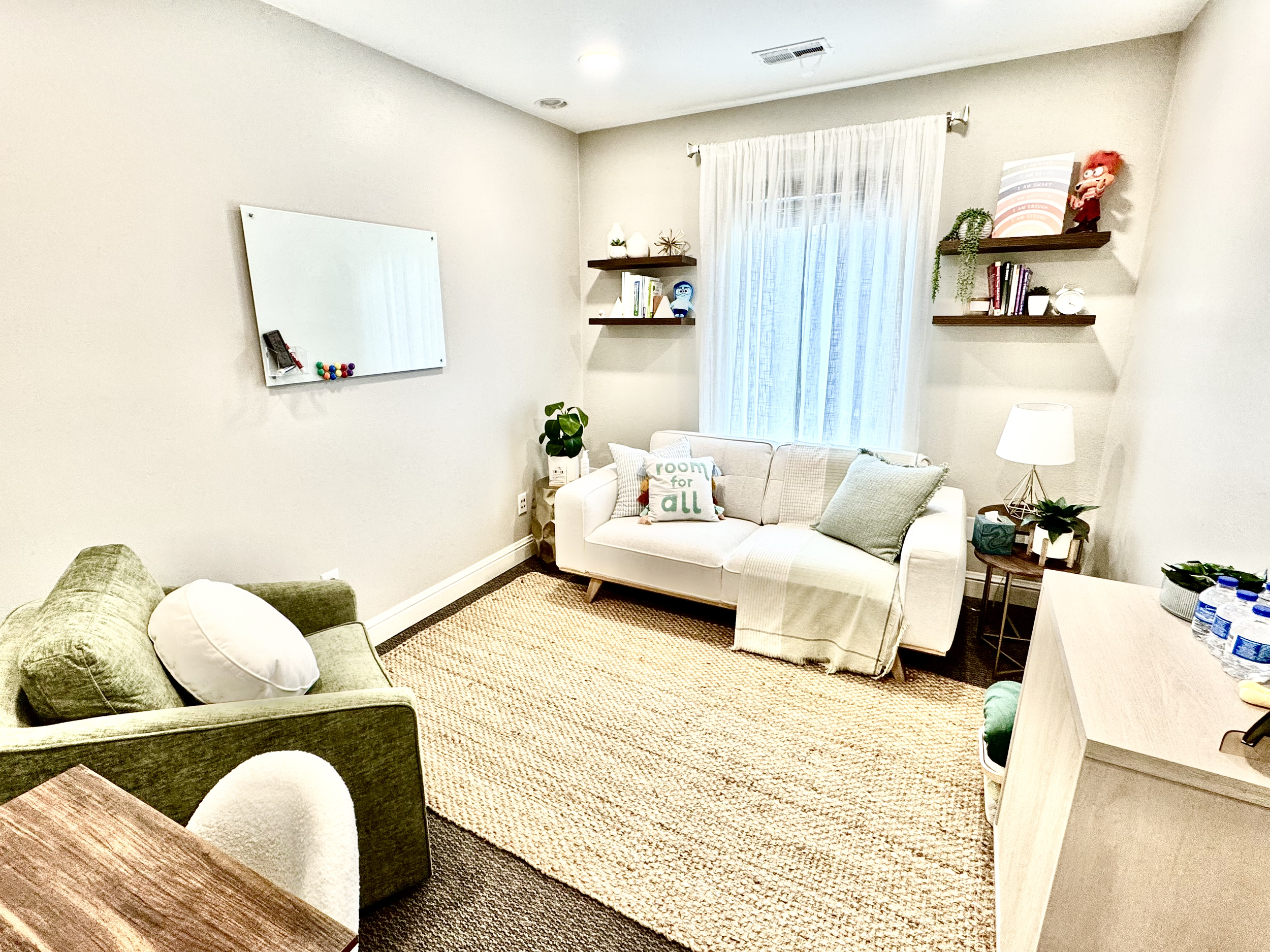 Cozy living room with a beige carpet, a white sofa with pillows, a green armchair, a wooden desk, wall shelves with books and decor, and a window with sheer curtains.
