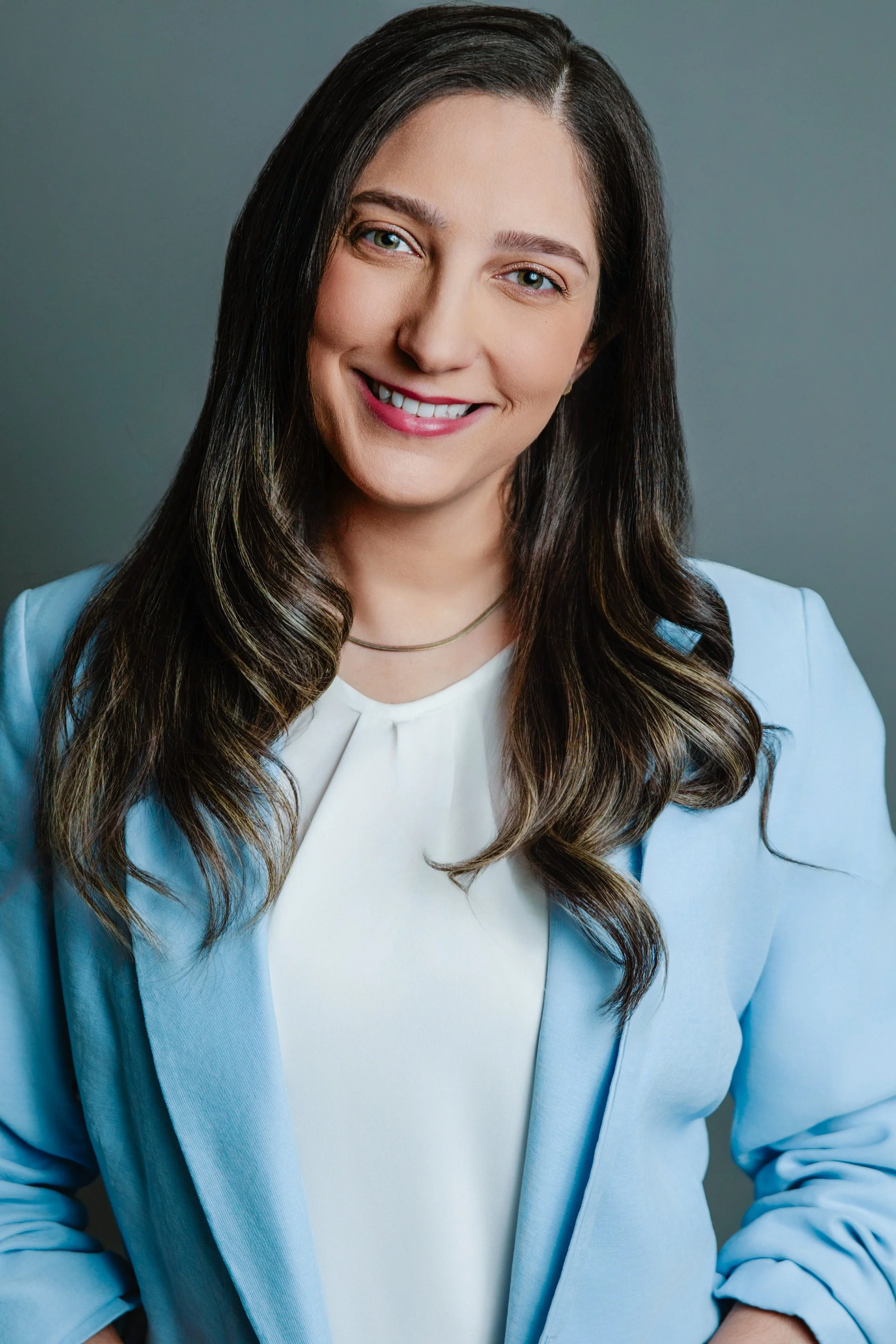 Portrait of a woman with long dark hair, smiling, wearing a light blue blazer over a cream top, with a plain gray background.