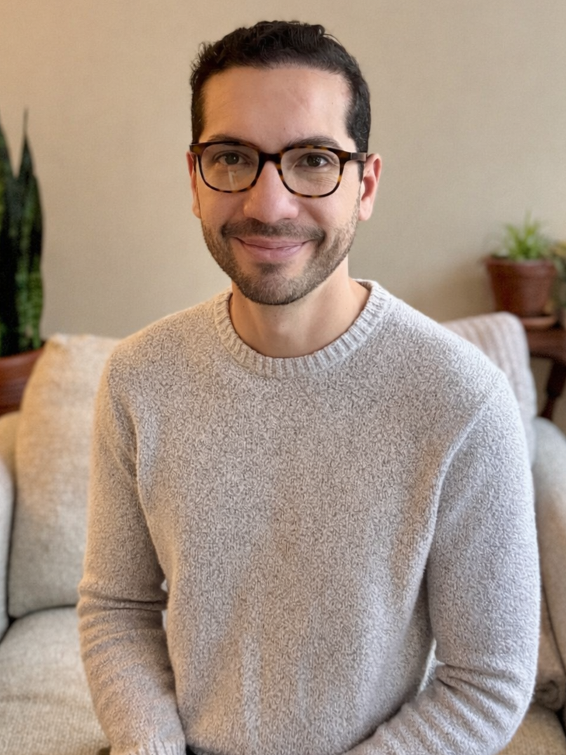 A man with dark hair, glasses, and a beard, smiling, standing outdoors with blurred fall foliage in the background.