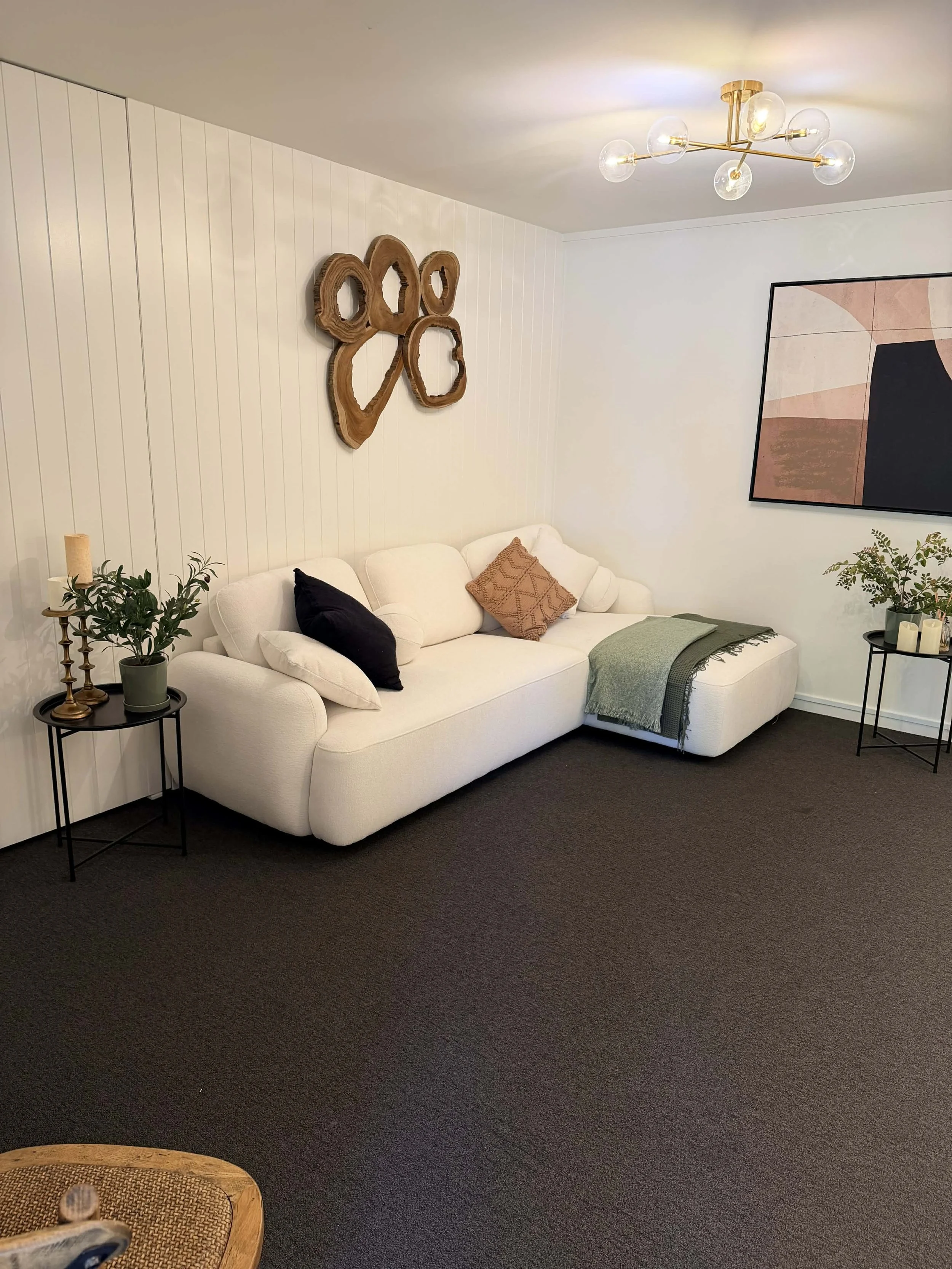 Visitor room with white sectional sofa with black, beige, and patterned pillows, dark carpet, wall art, wood wall decor, side tables with plants and candles, and a modern ceiling light fixture.
