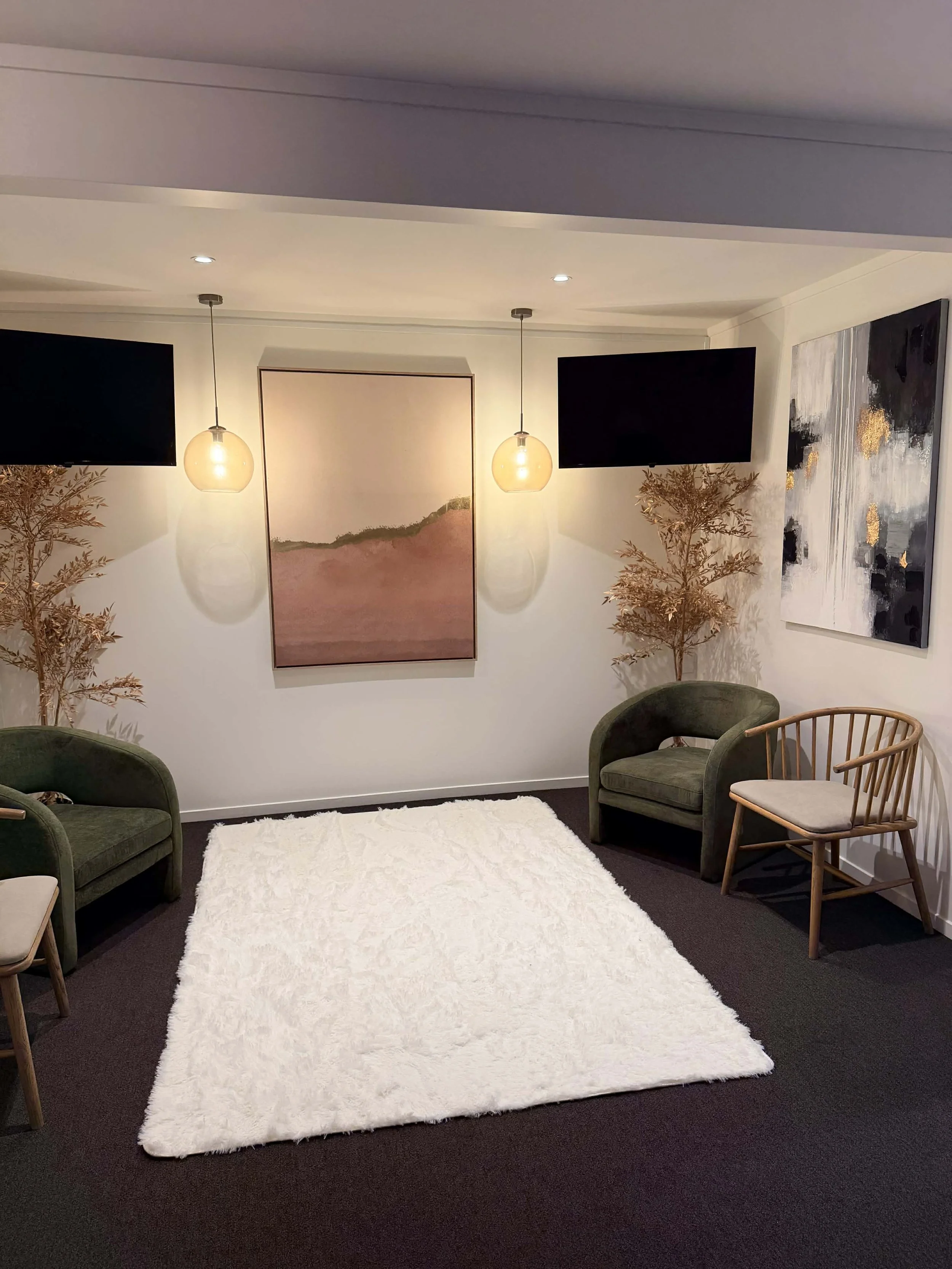 Visitor room with two green armchairs, a wooden chair, a white rug, artwork on the walls, two hanging pendant lights, and decorative trees.