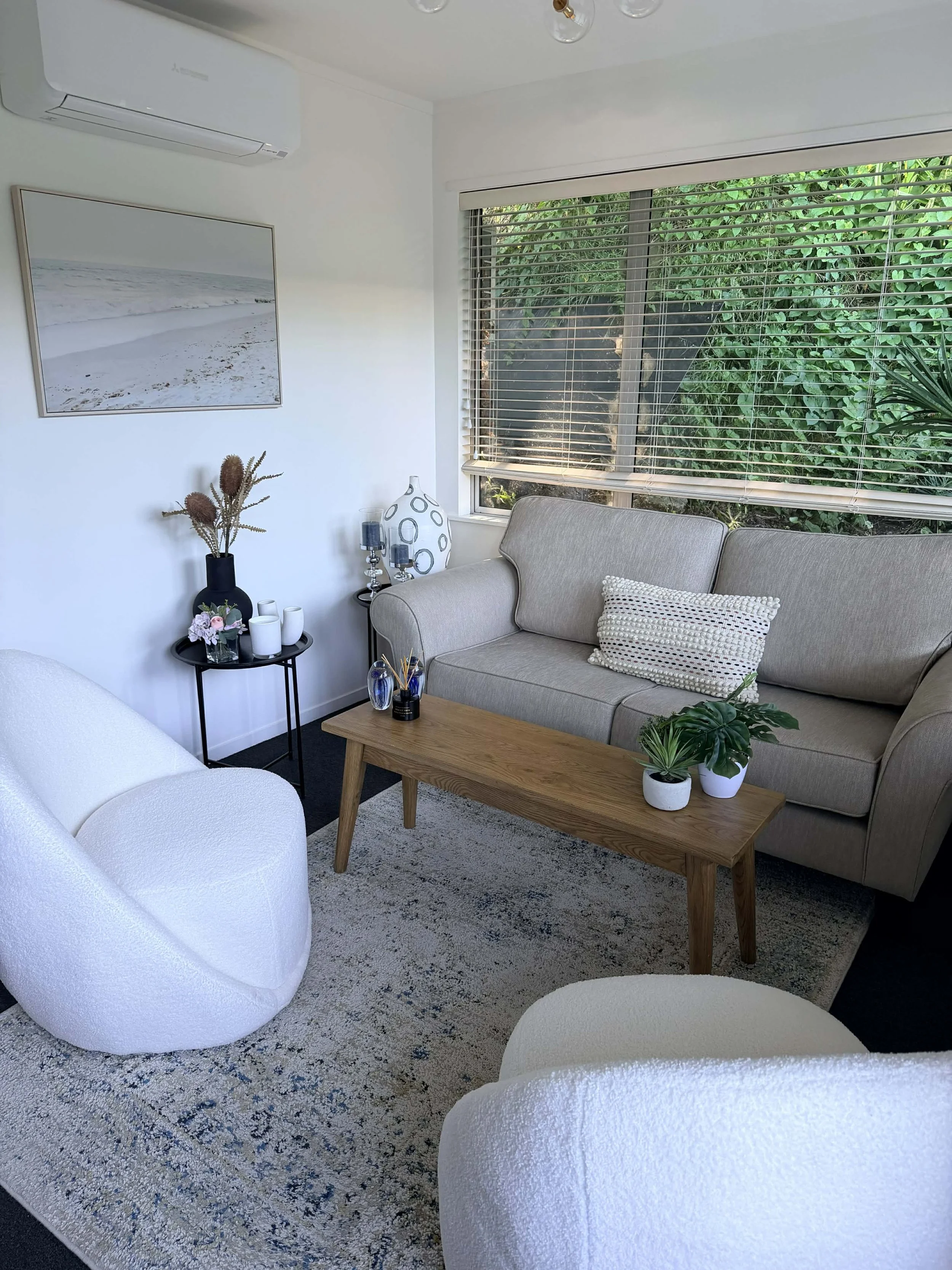 Visitor room with beige couch, white armchair, wooden coffee table, decorative vases, potted plants, window with blinds, and framed beach photograph.