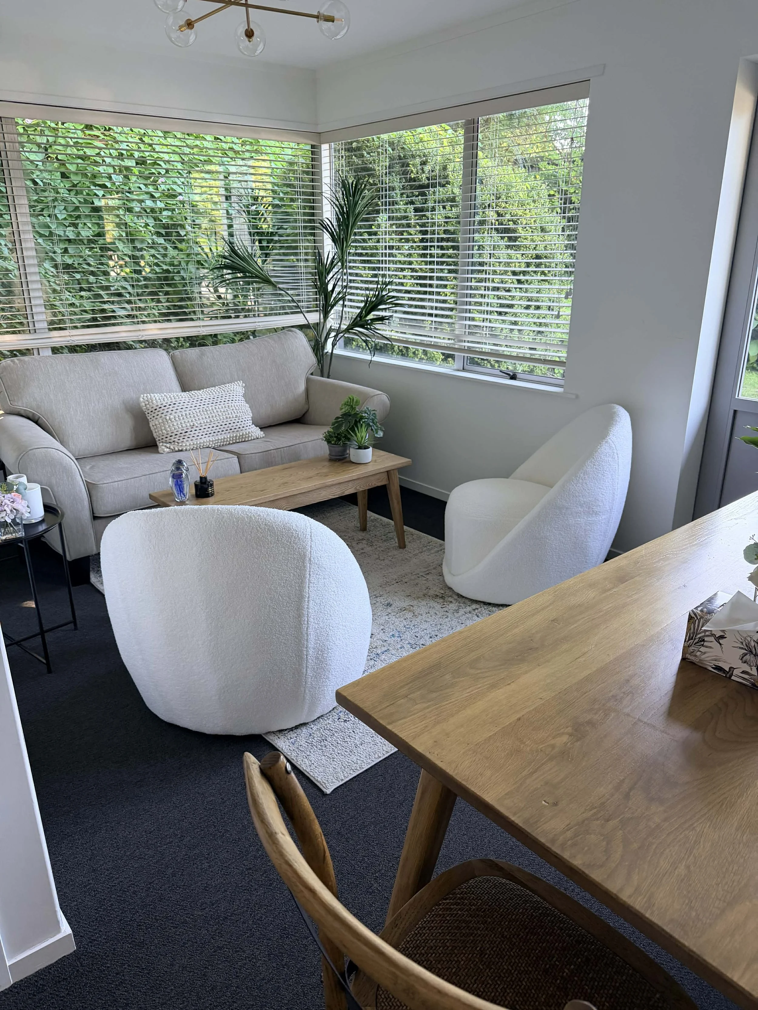 Visitor room with large windows, beige sofa, white armchair, round white chair, wooden coffee table, indoor plants, and a black side table with plants and decor.