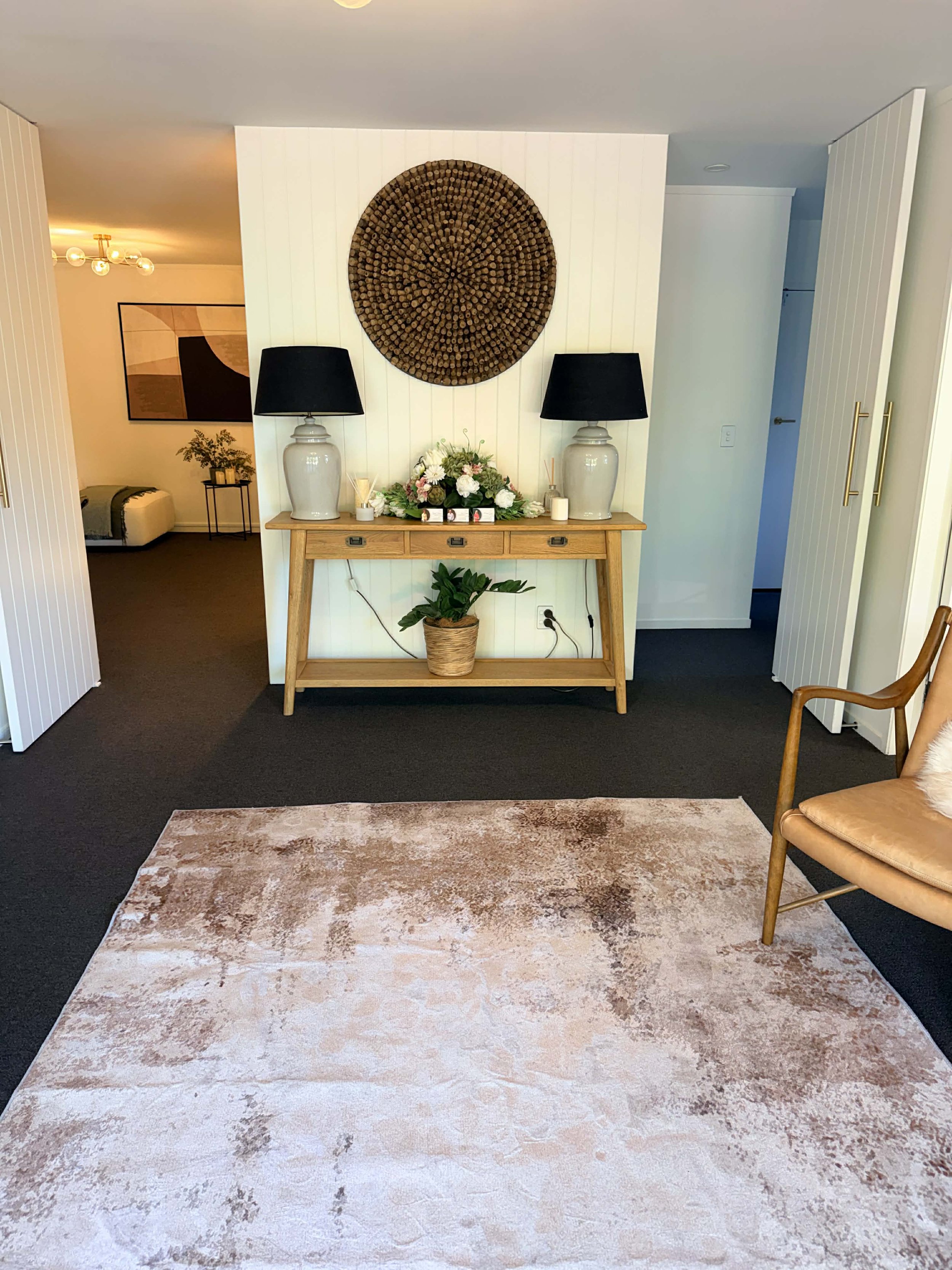 Visitor room with a wooden console table, two large lamps, and a flower arrangement, against a white paneled wall with a circular decorative wall hanging made of wood slices, a pink and beige area rug, and a tan armchair.