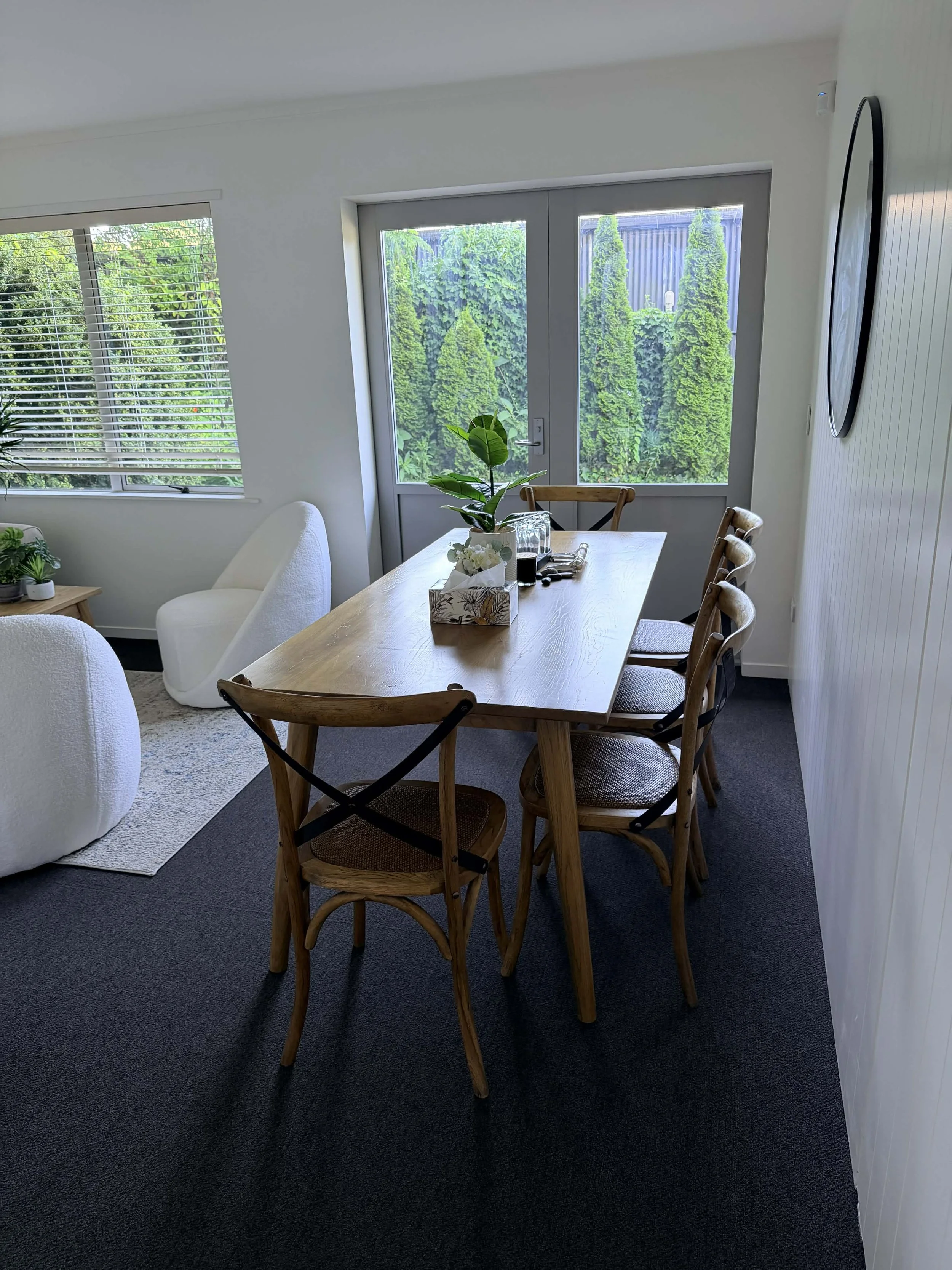 Visitor room with a wooden table, six chairs, a potted plant, and large windows showing greenery outside.