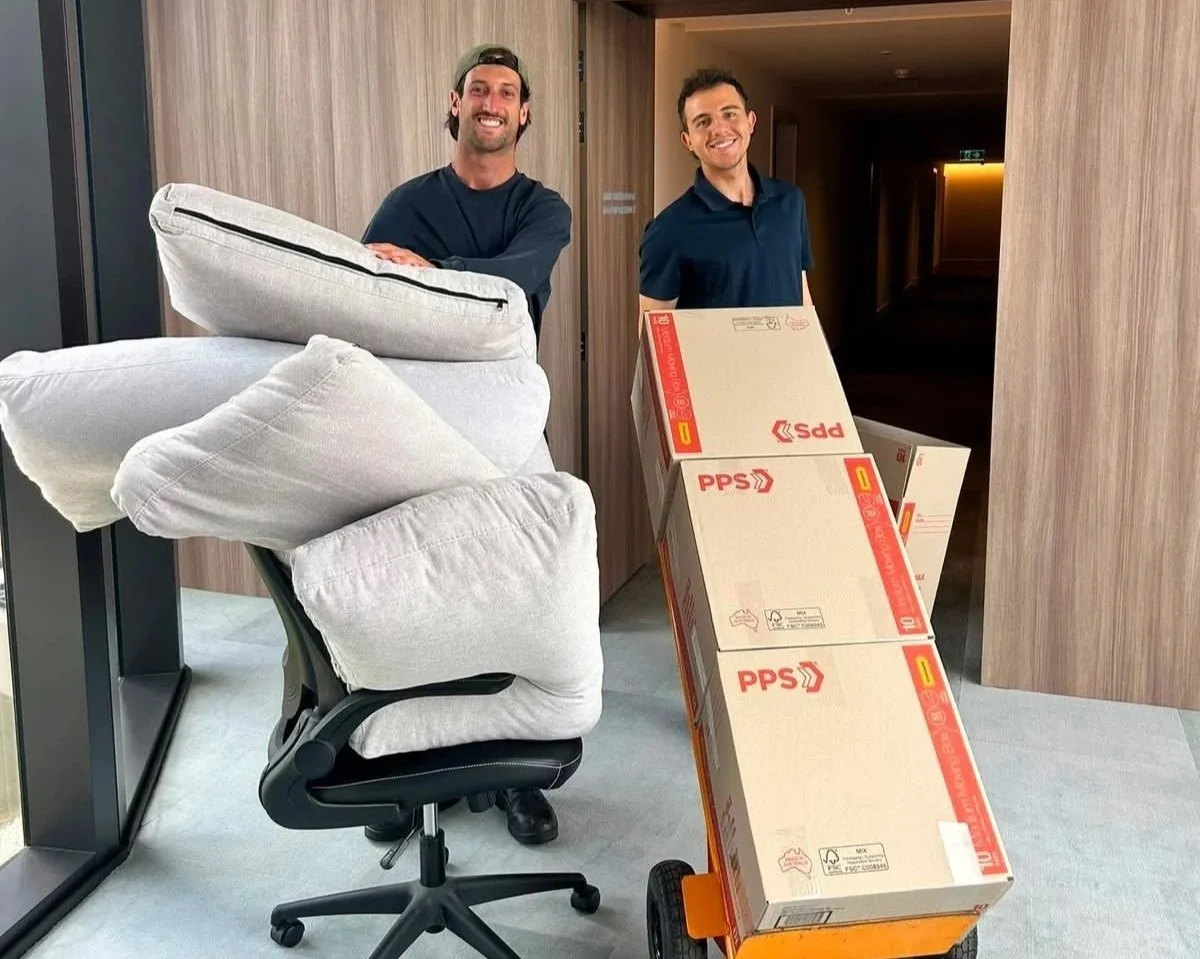 Two smiling men, one with a beard and the other clean-shaven, are moving boxes and pillows in an indoor hallway. They are standing behind a hand truck loaded with large cardboard boxes labeled PPS.