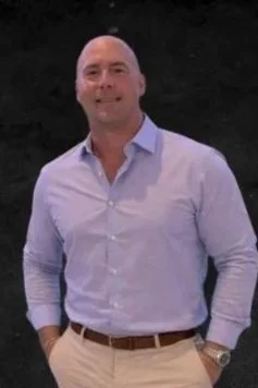 A man with a shaved head wearing a light blue button-up shirt and beige pants, smiling outdoors against a dark background.