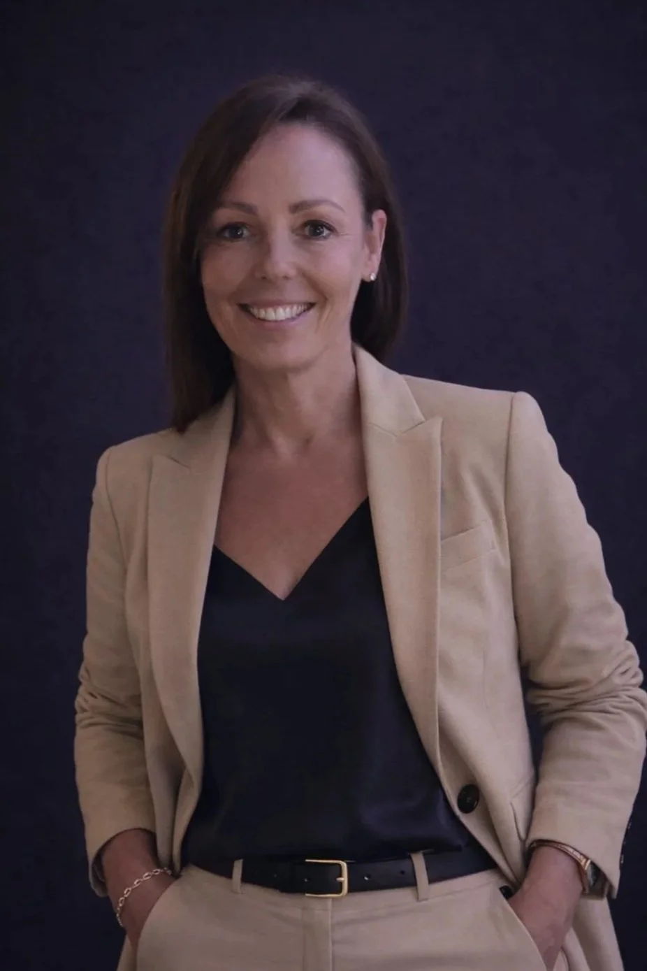 A woman with shoulder-length brown hair wearing a beige blazer, black top, and beige pants, smiling against a dark background.