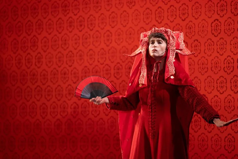 A woman dressed in elaborate red costume with a headpiece, holding a black and red fan, standing against a red patterned backdrop.