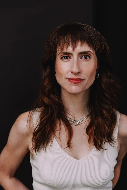Portrait of a woman with shoulder-length brown hair, wearing a white sleeveless top and layered necklaces, standing against a dark background.