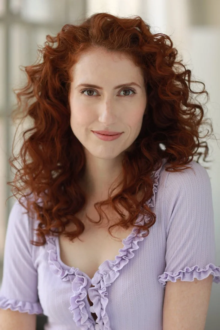 Close-up of a woman with curly red hair and blue eyes, wearing a light purple top with ruffled edges, smiling softly indoors.