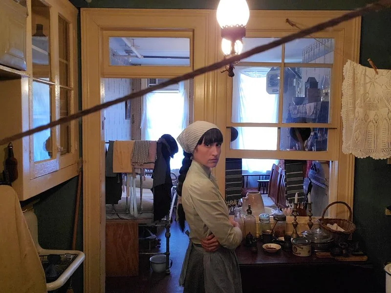 Young woman dressed in vintage clothing and a white headscarf standing in a kitchen, looking at the camera, with a dining area and laundry visible in the background.